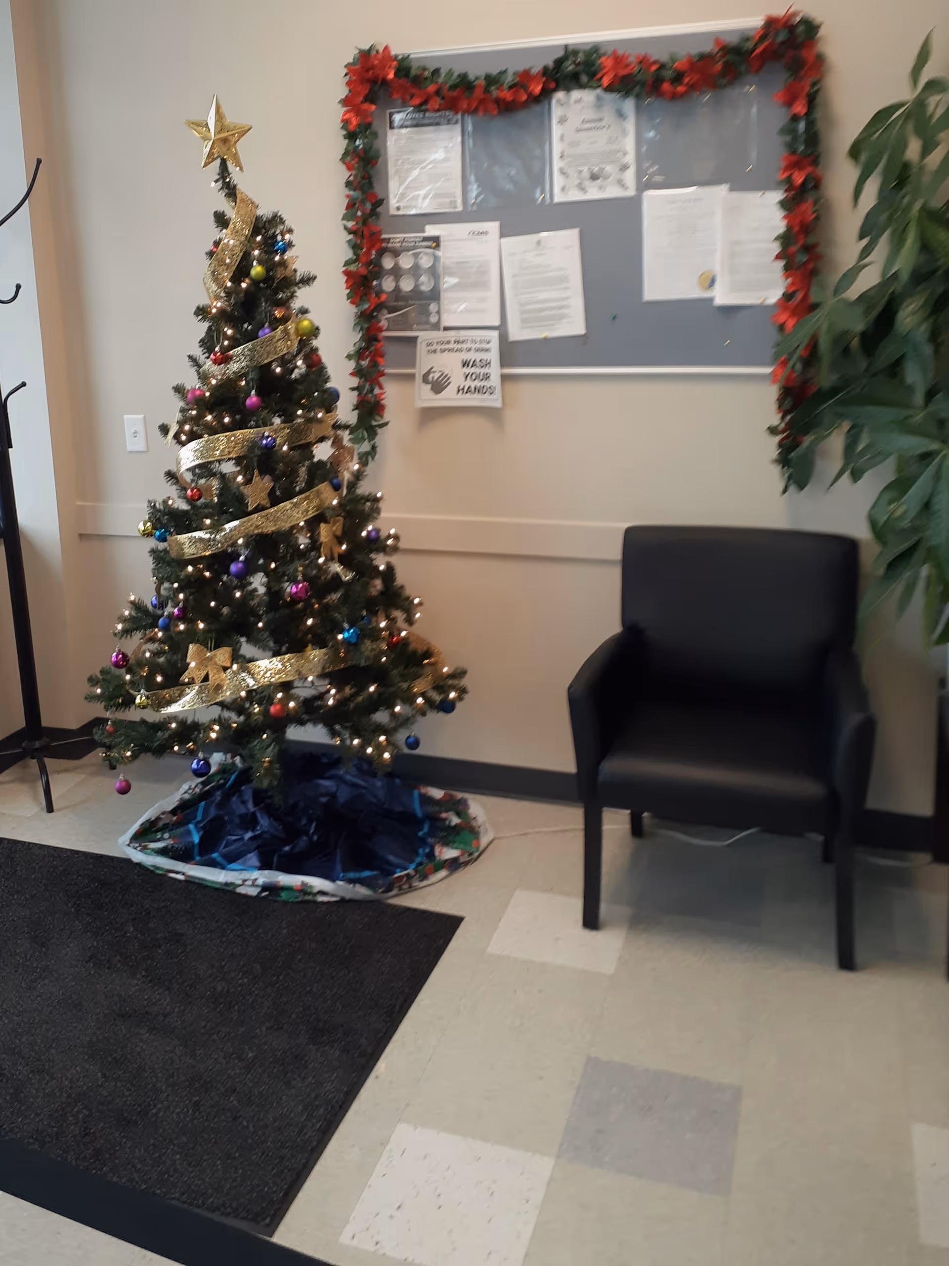 A decorated Christmas tree beside a black chair and bulletin board in a facility lobby.