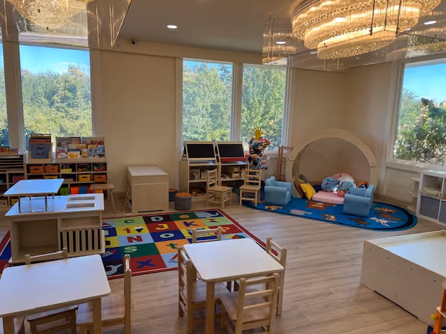 Bright interior playroom with small tables, shelves, a colorful rug, and a cozy play tent.