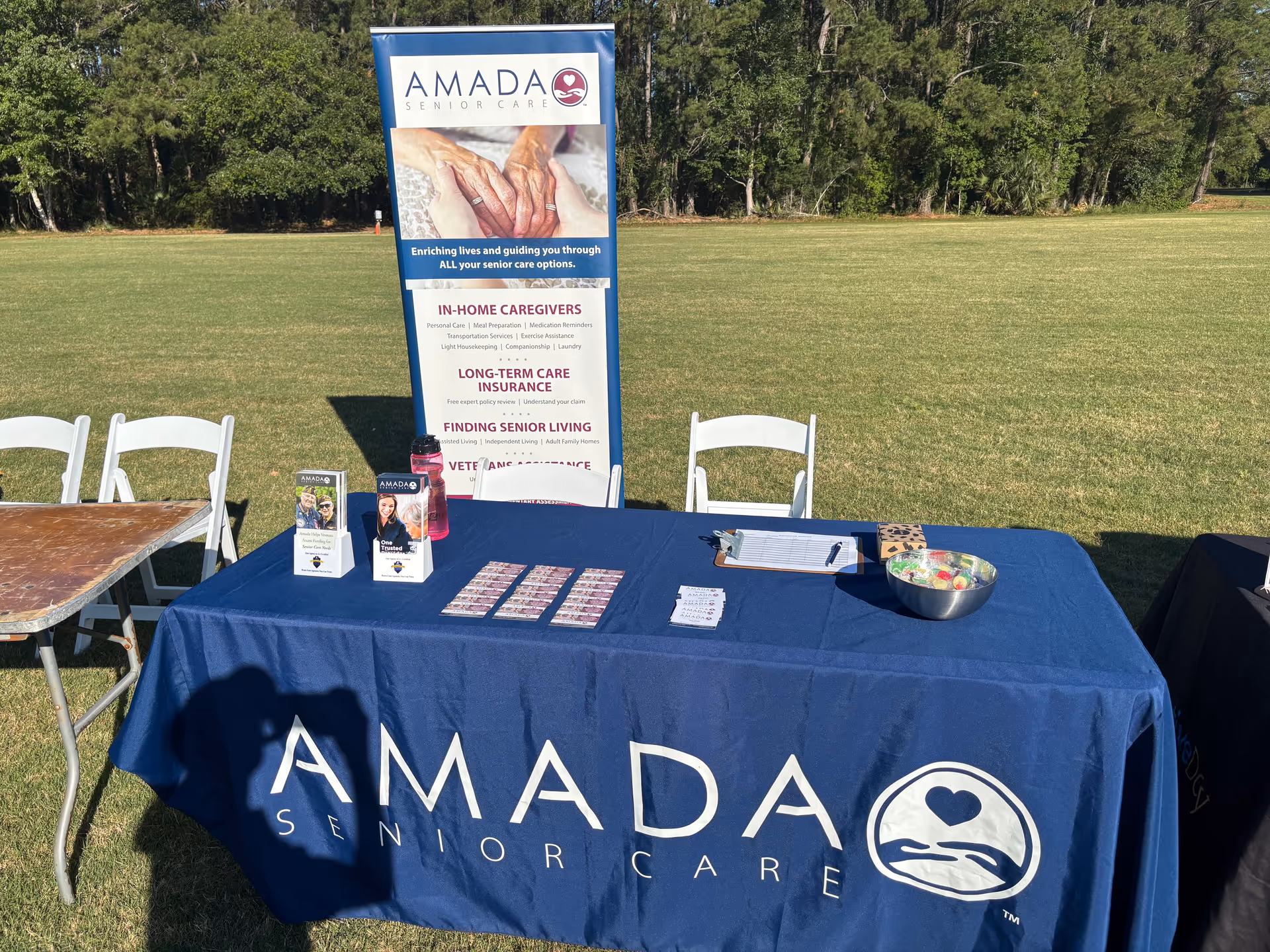 Outdoor promotional booth for Amada Senior Care with blue tablecloth, banner, and white chairs on grass.