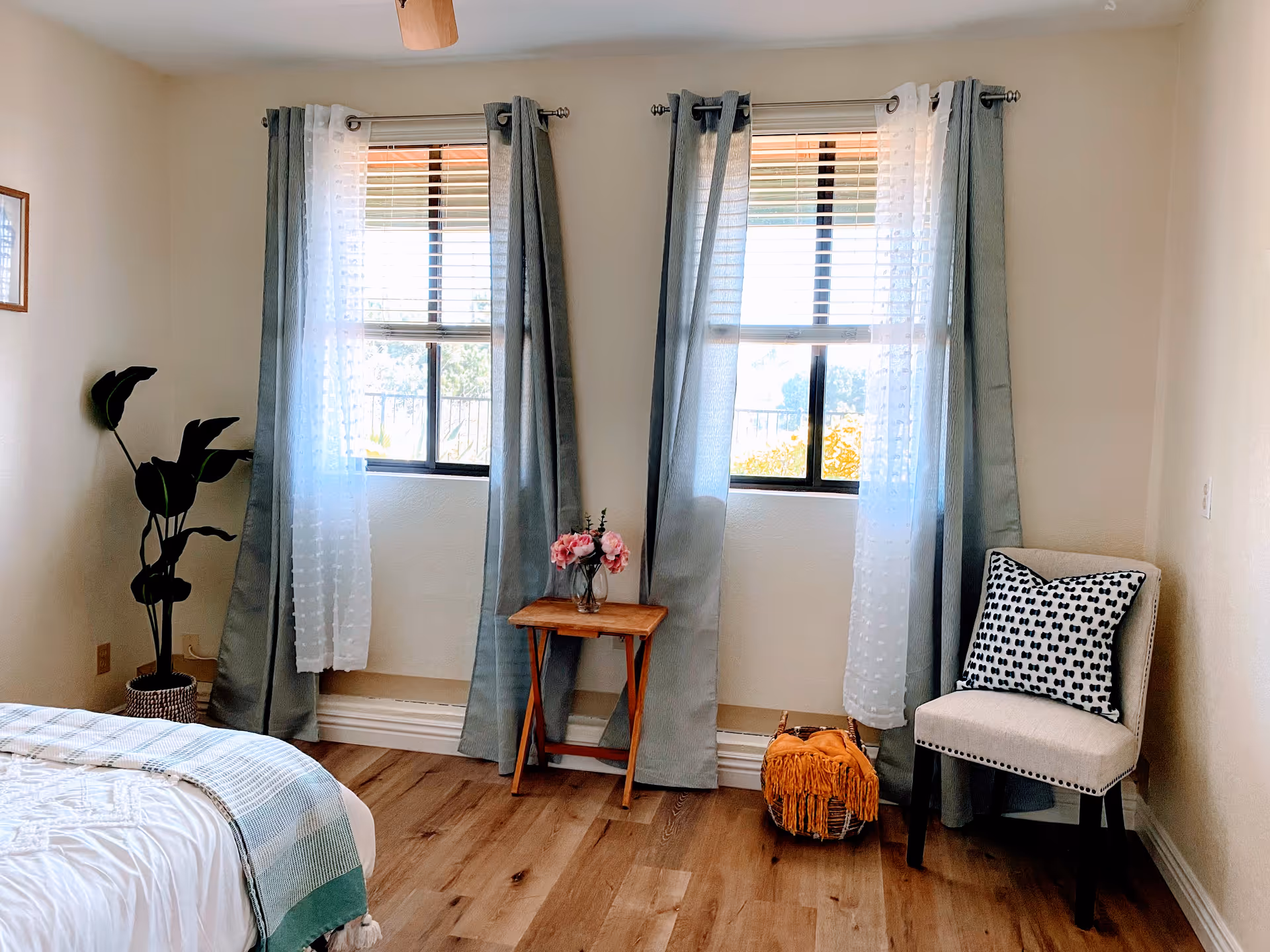 Bright bedroom with two windows, curtains, a table with flowers, chair, and visible bed.