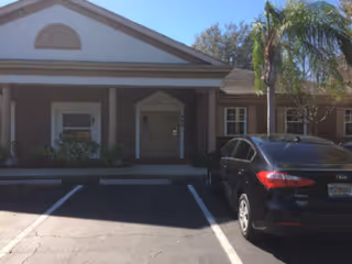 Front exterior of a brick senior facility with palm trees and a parked car.