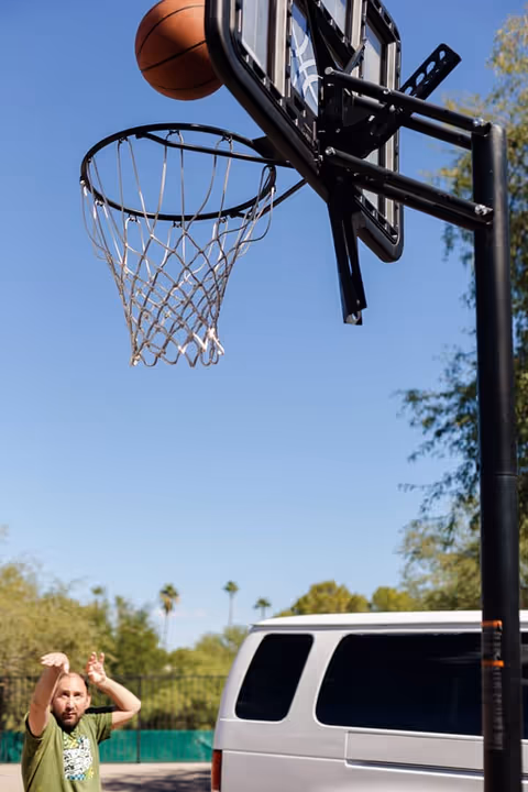 Basketball hoop with ball in mid-air and a man near a white van outdoors.