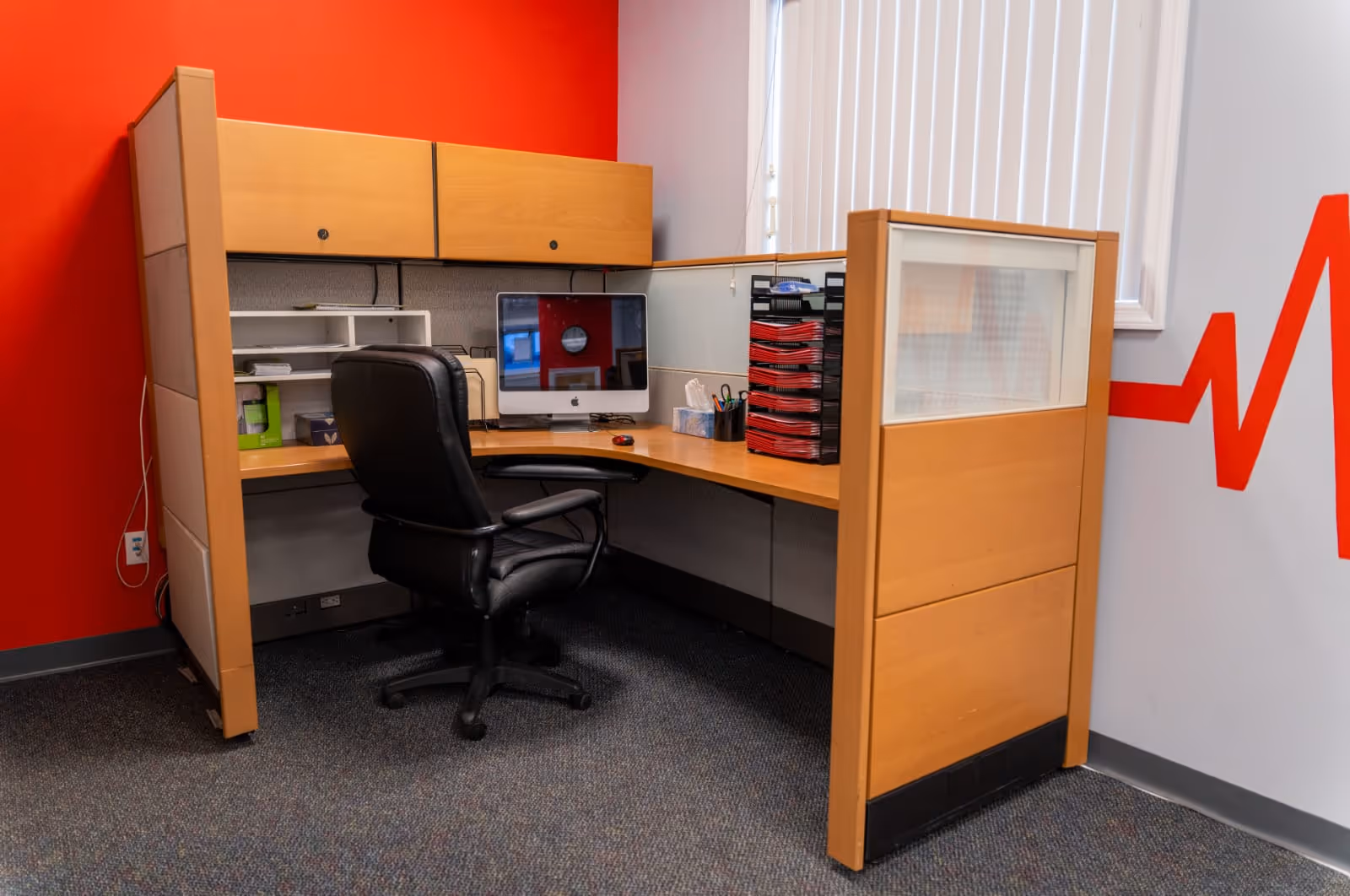 A compact office cubicle with a desk, chair, computer, and red wall.