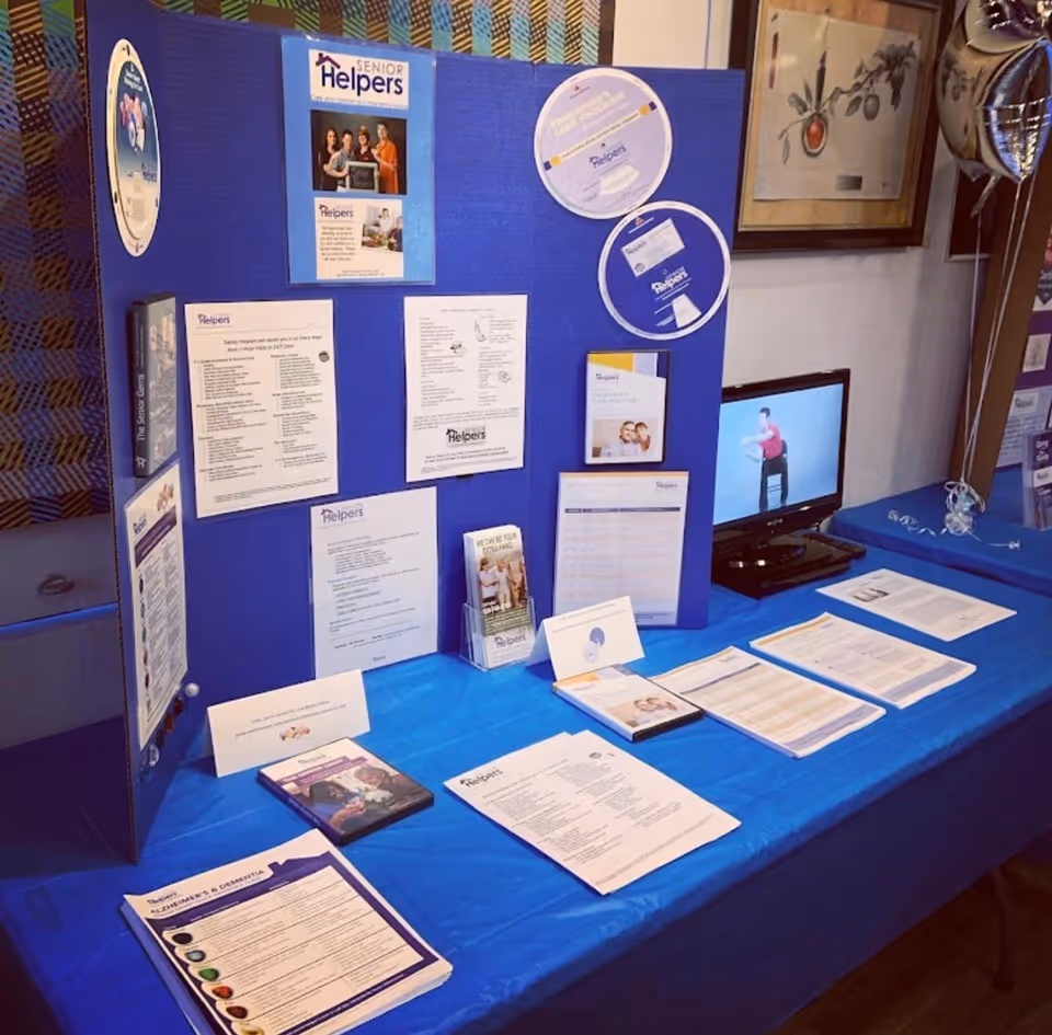 Blue display board with Senior Helpers brochures and a laptop on a blue table indoors.