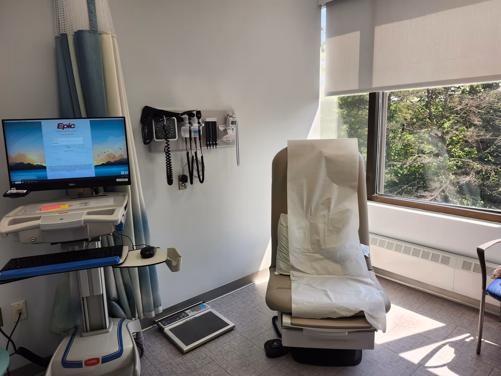 Medical examination room featuring a chair with paper sheet, monitor, and wall-mounted devices.