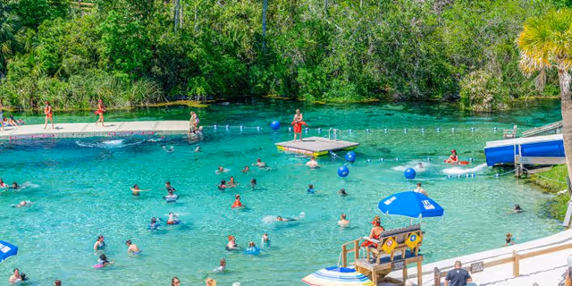 Outdoor swimming area with numerous swimmers in turquoise water and deck seating.