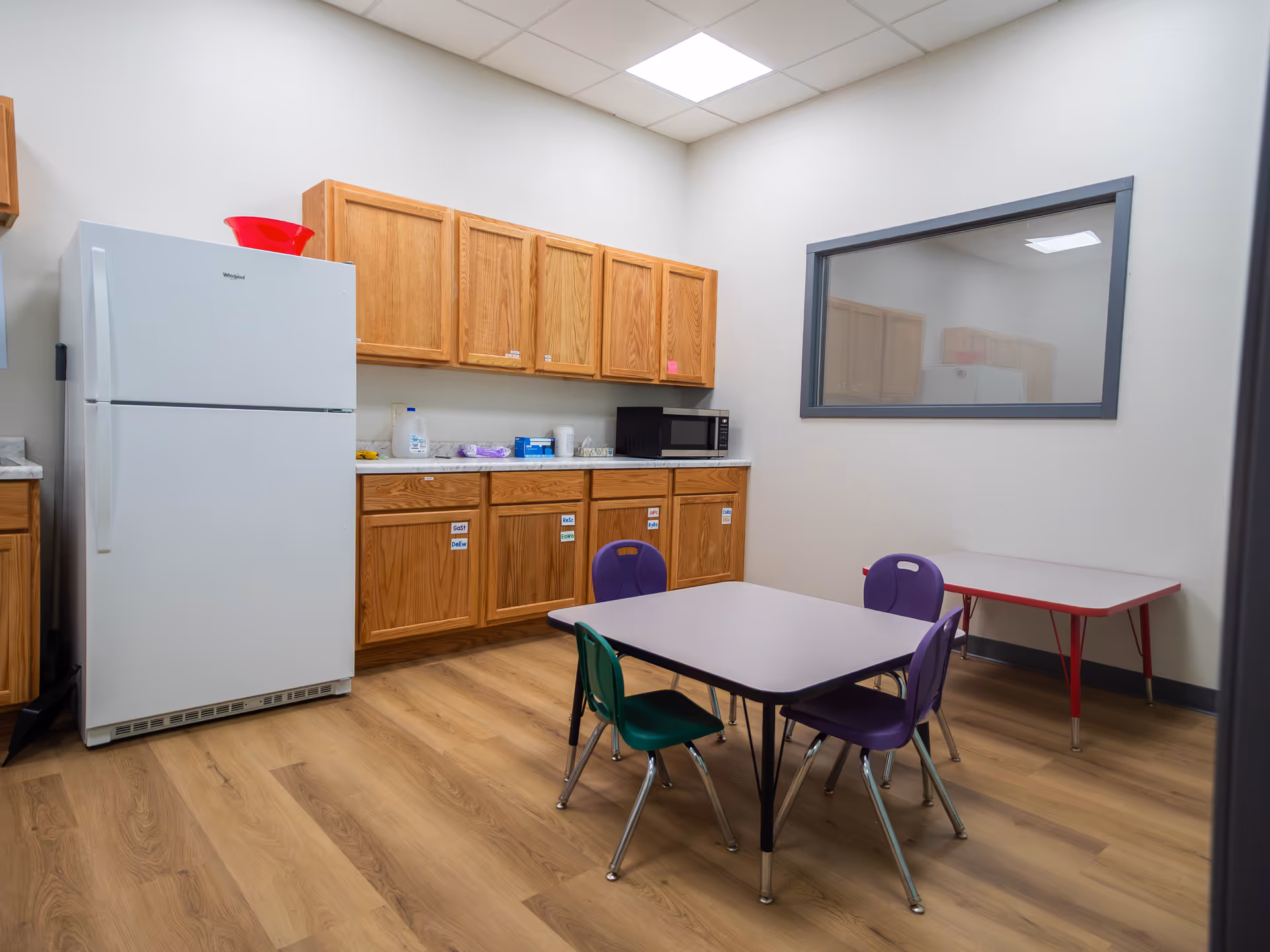 A small kitchen area with wooden cabinets, fridge, microwave, and a table with colorful chairs.