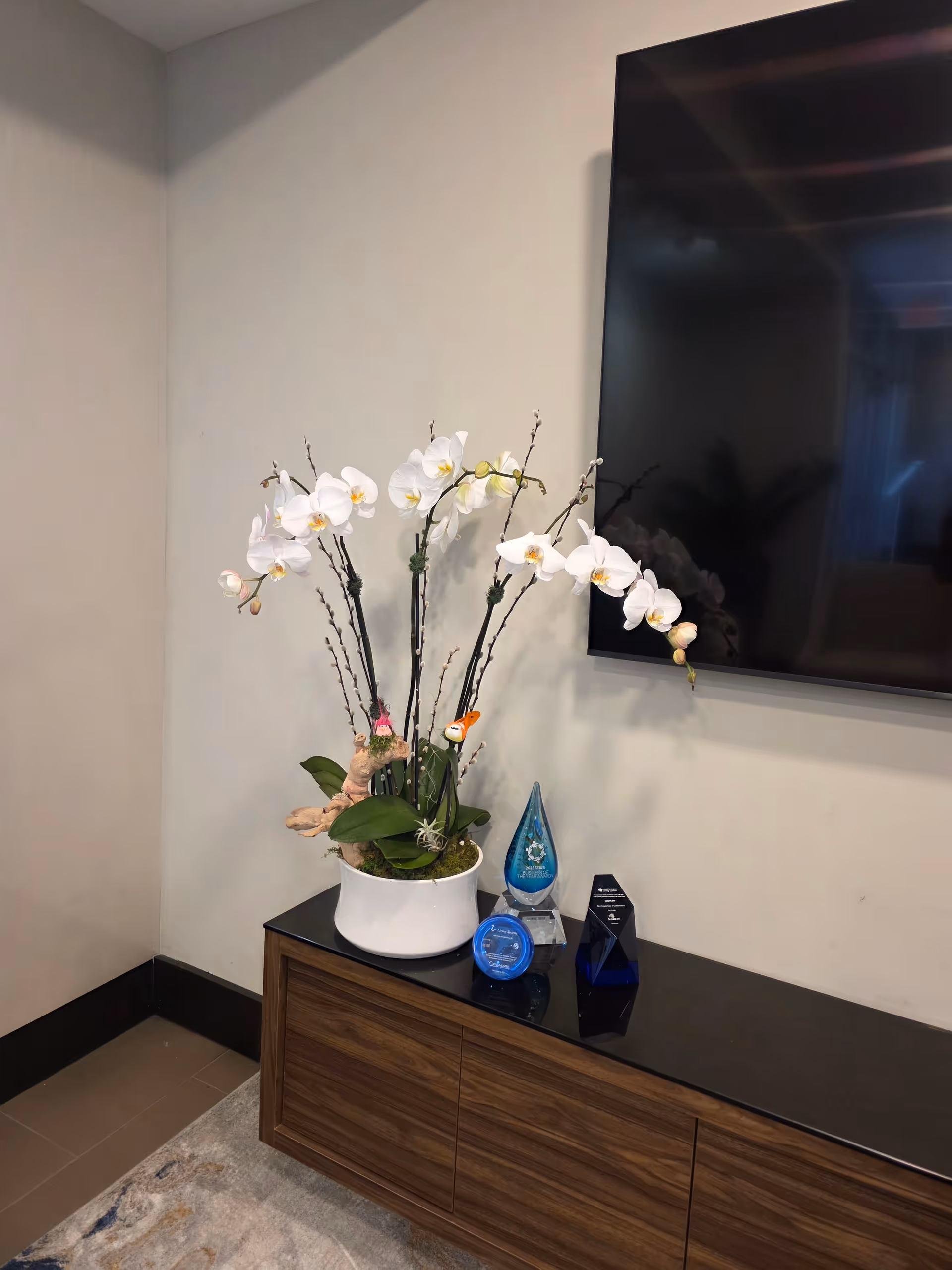 White orchid arrangement on a dark wooden console with a wall-mounted TV.