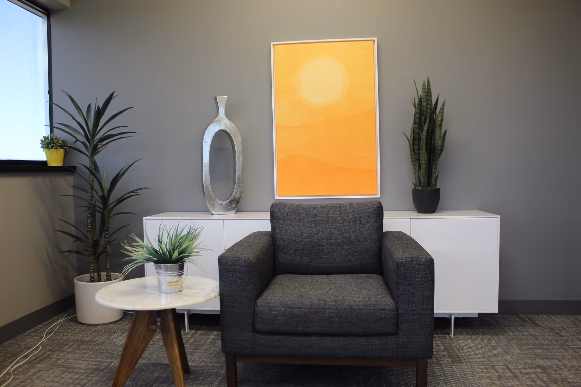 Modern lounge with gray armchair, plants, a round table, and orange artwork.