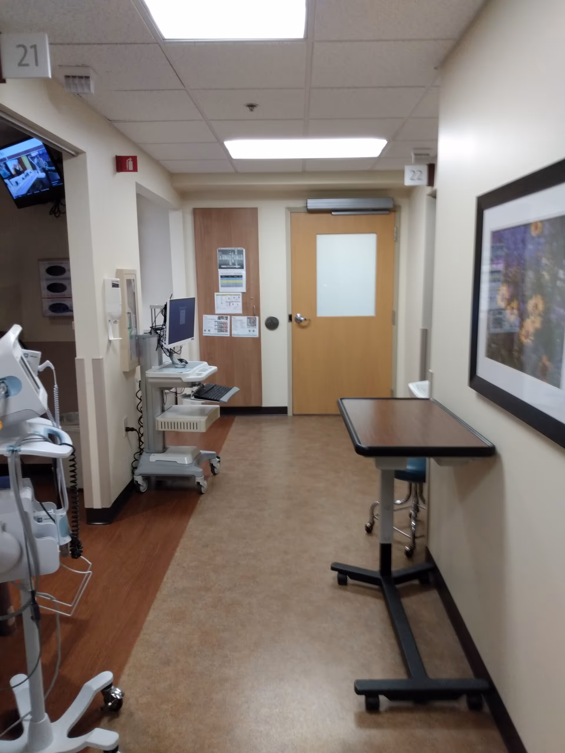 Interior hospital corridor with medical equipment carts, doors, and a standing table along the wall.