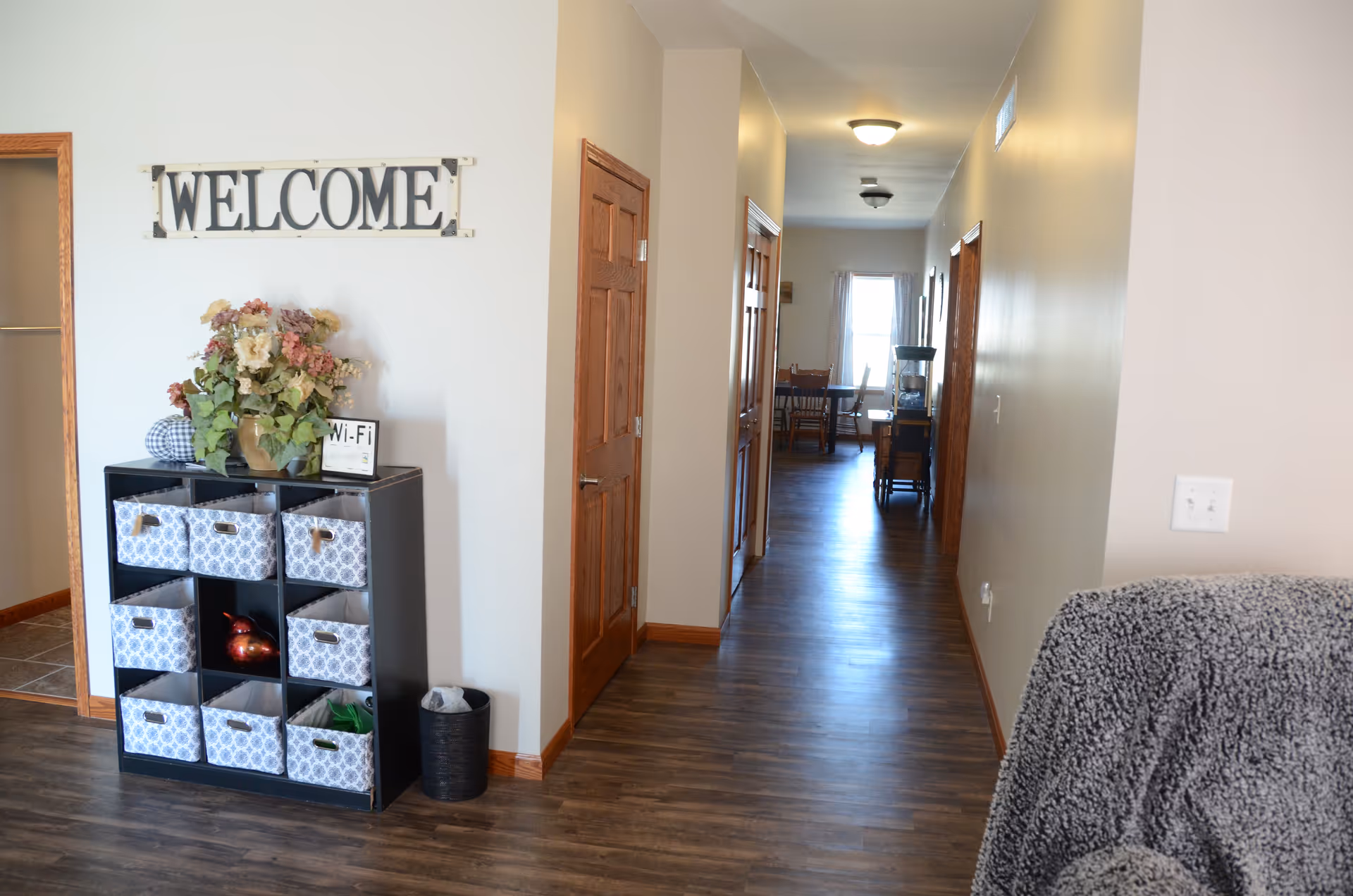 Indoor hallway in a senior care facility with a welcome sign, cubbies, plant, and couch.