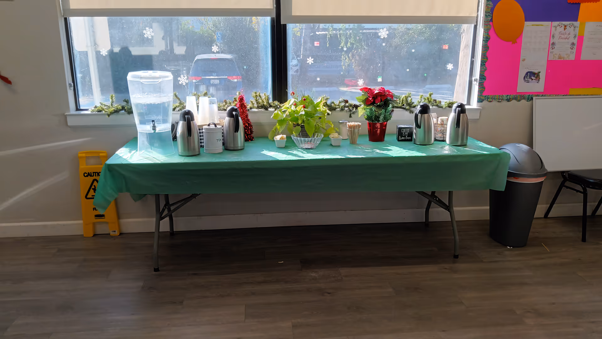A table with a green tablecloth holds coffee kettles, water dispenser, cups, and plants.