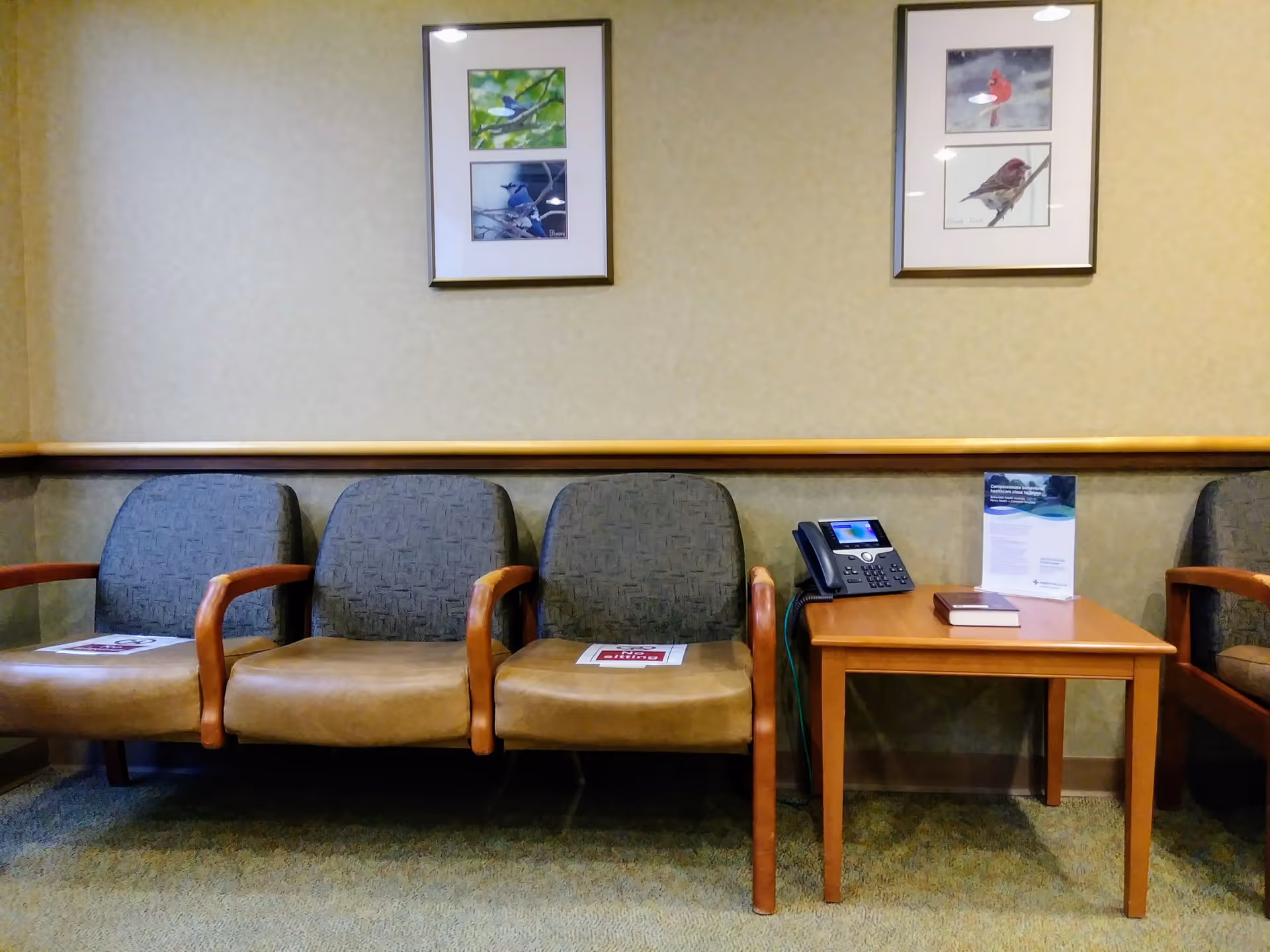 Waiting area with cushioned chairs, a wooden table, phone, and framed art on beige walls.