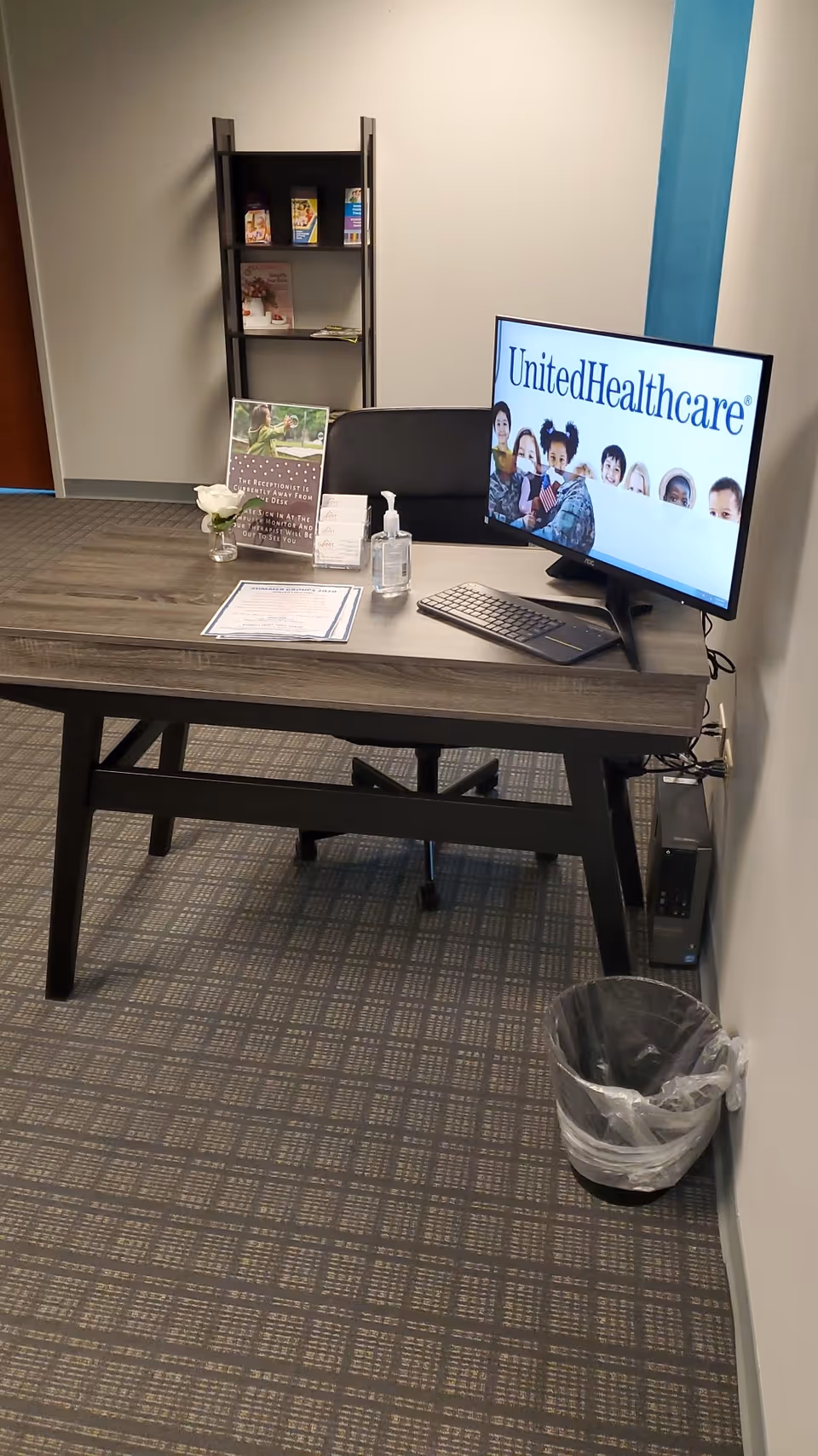 Reception desk with computer monitor, brochures, hand sanitizer, and trash can in an office area.