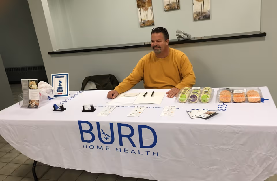 A man sits at a Burd Home Health booth table indoors with materials.