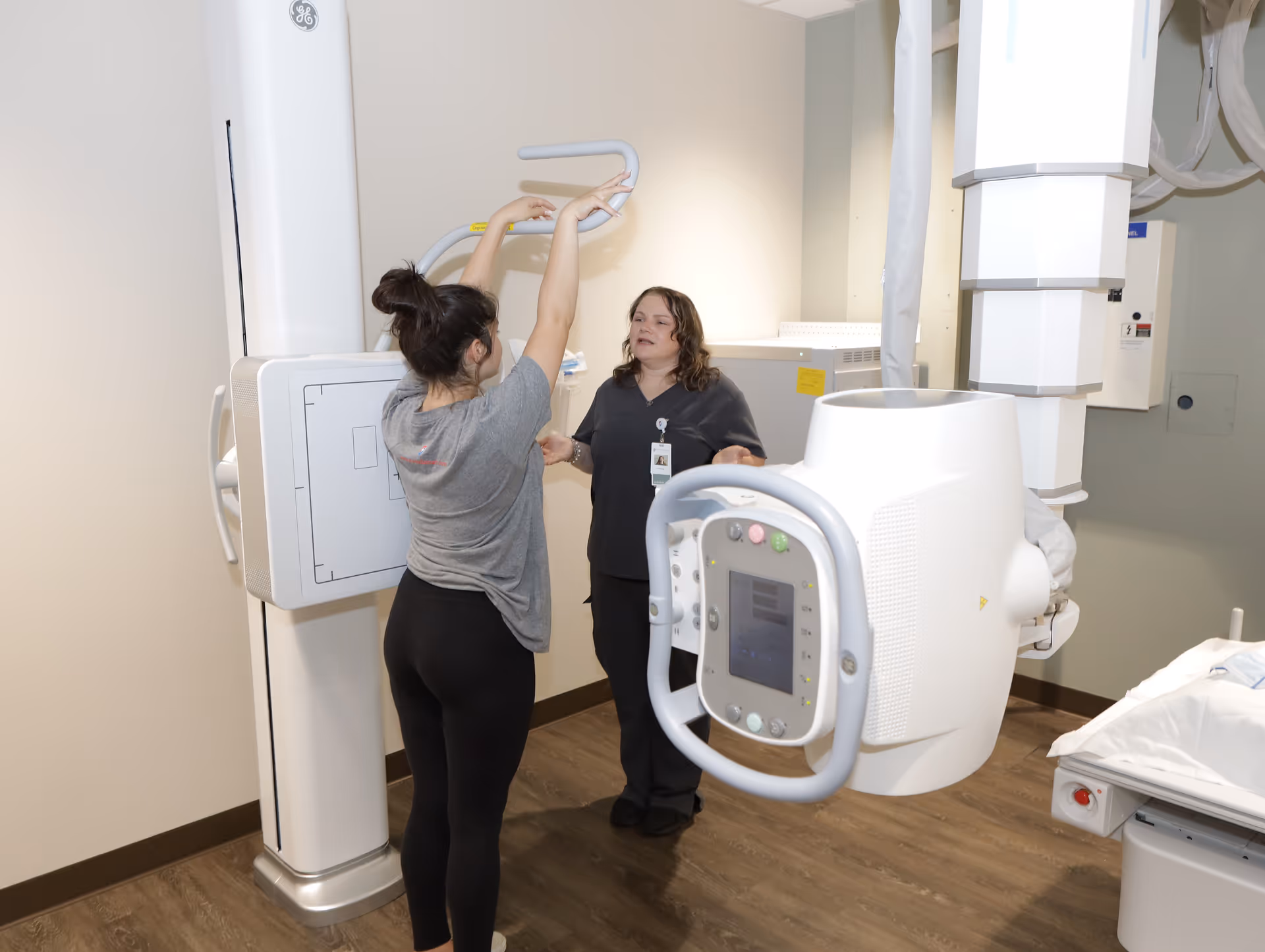 Two healthcare workers operate a large radiology machine in a hospital imaging room.