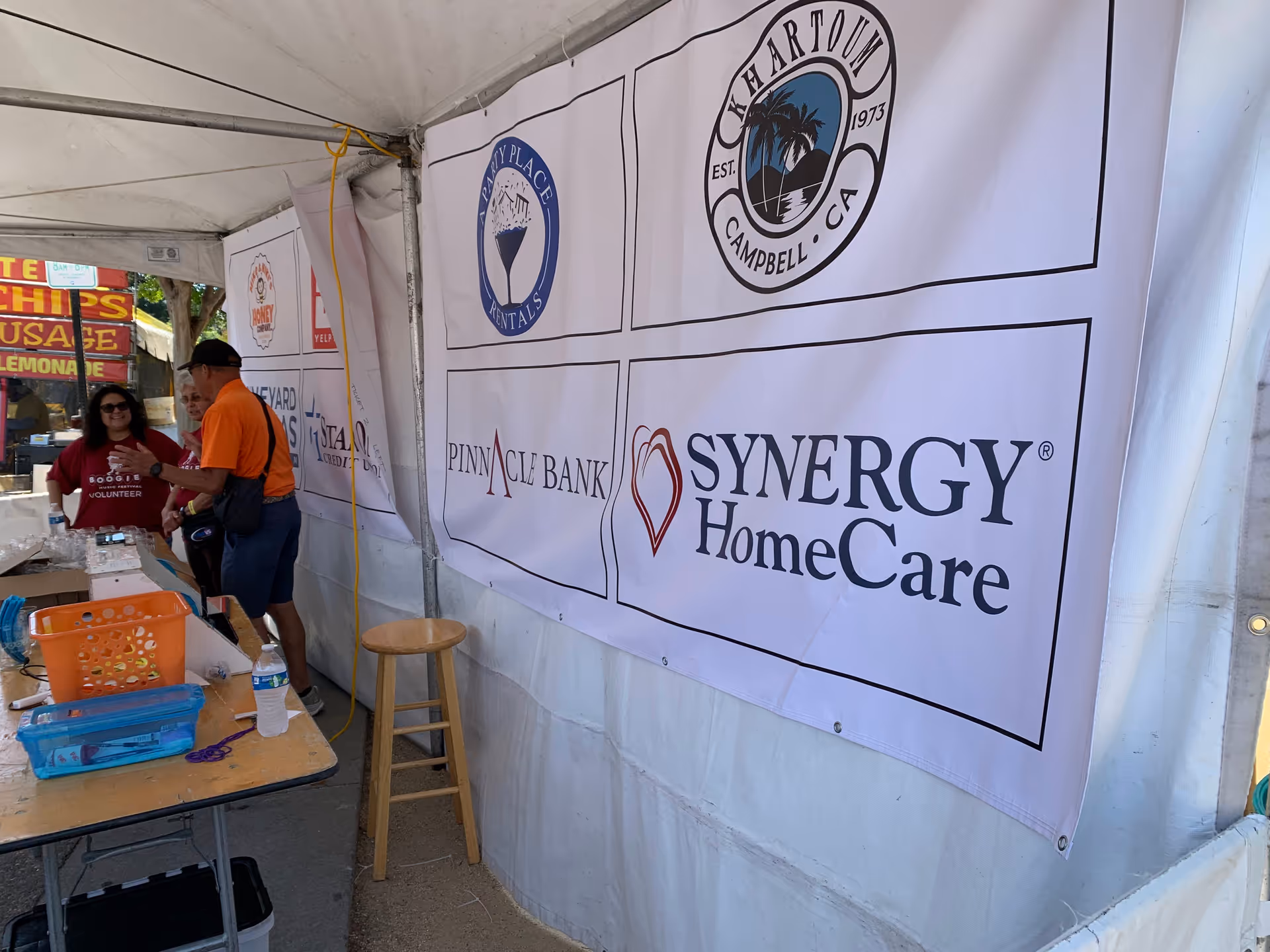 Outdoor event tent with Synergy HomeCare banners and volunteers at a registration table.