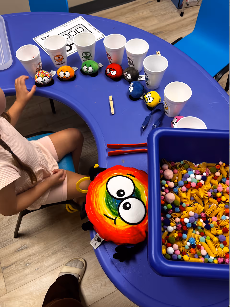 A child sits at a blue curved table with cups, colorful plush toys, and craft bins.