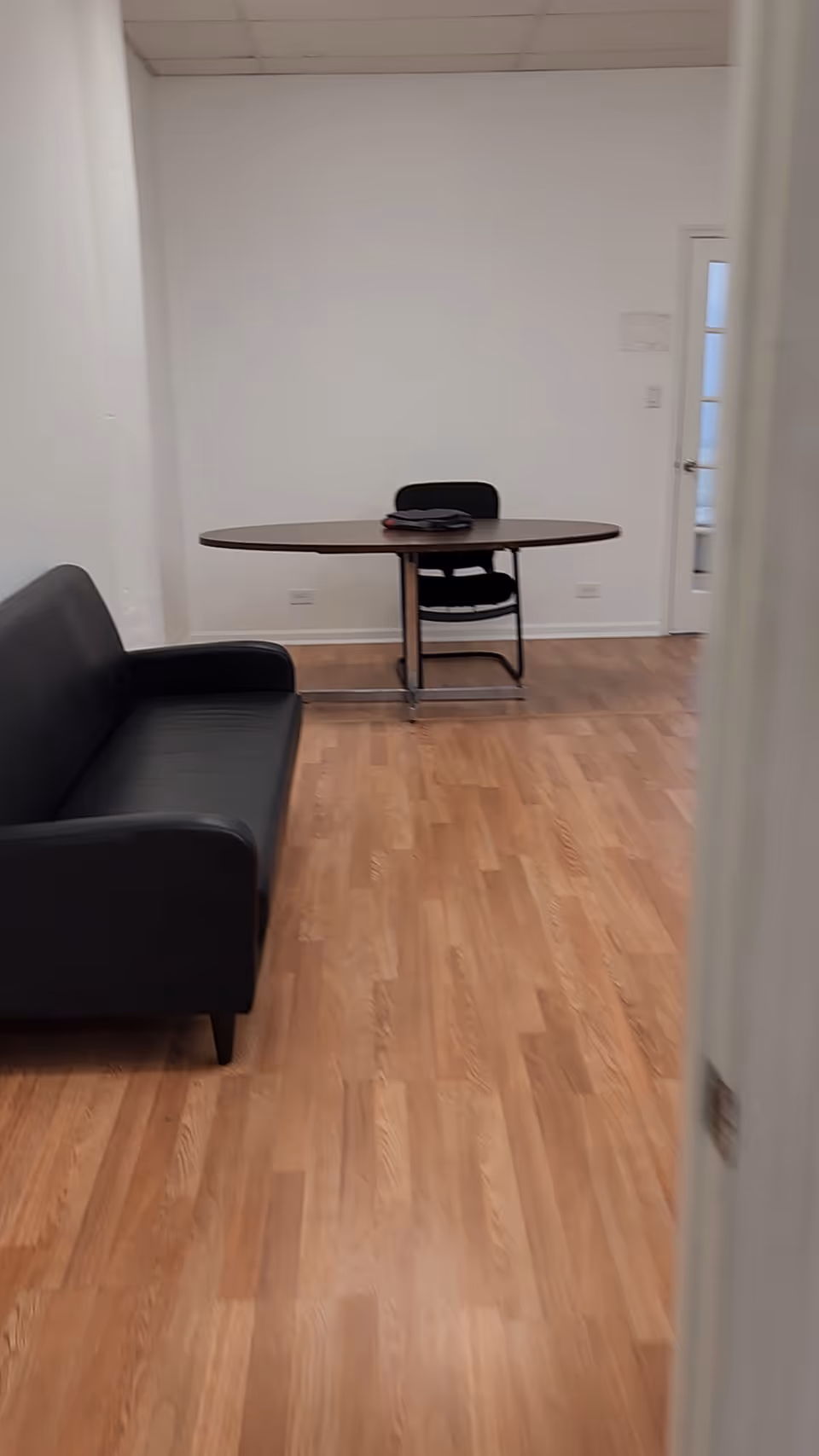 A small interior lounge with a black sofa, round table, chair, and wooden floor.