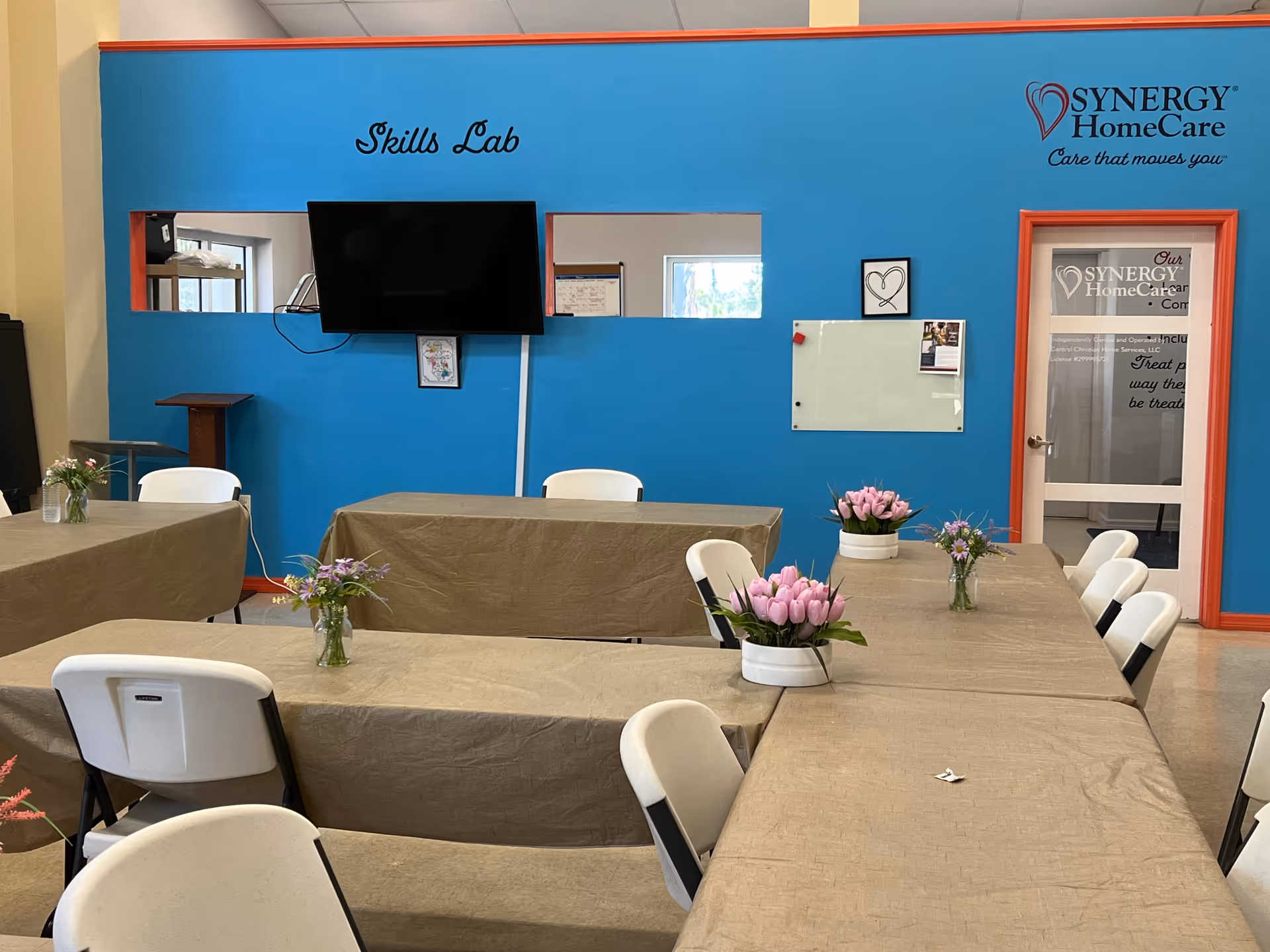 Blue-walled training area labeled 'Skills Lab' with TV, tables, chairs, and flowers.