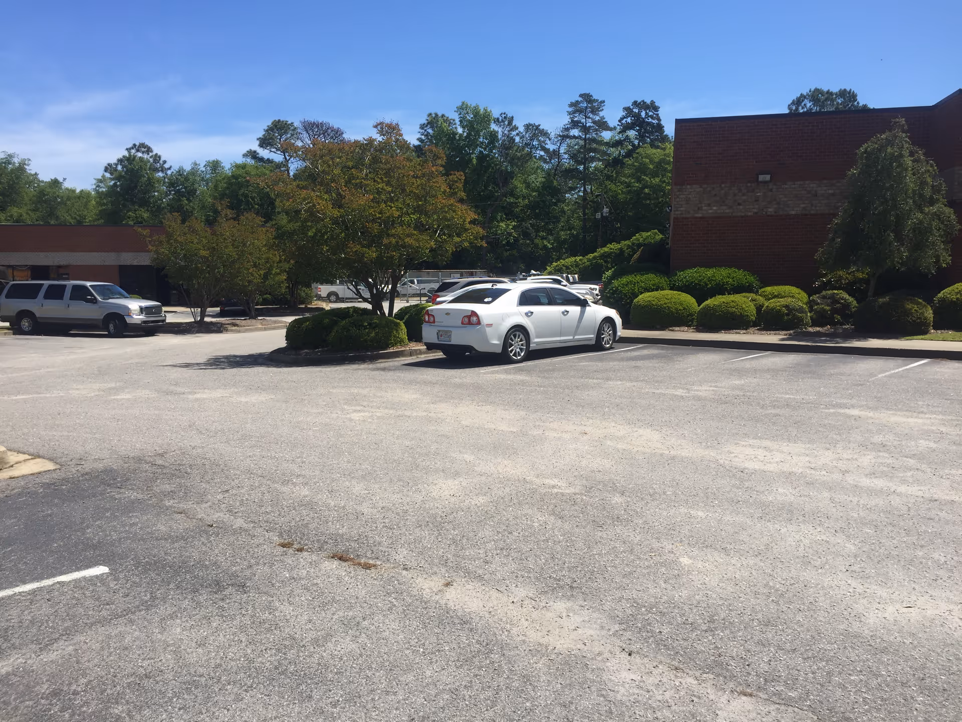 Parking lot in front of a brick building with trees, shrubs, and parked cars.