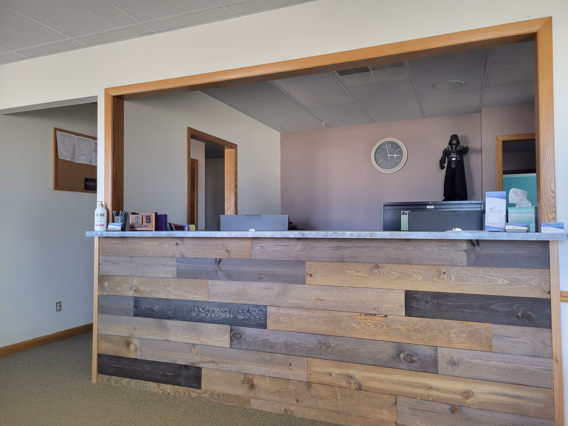 Reception desk with wooden paneling, clock on lavender wall, and a Darth Vader figurine.
