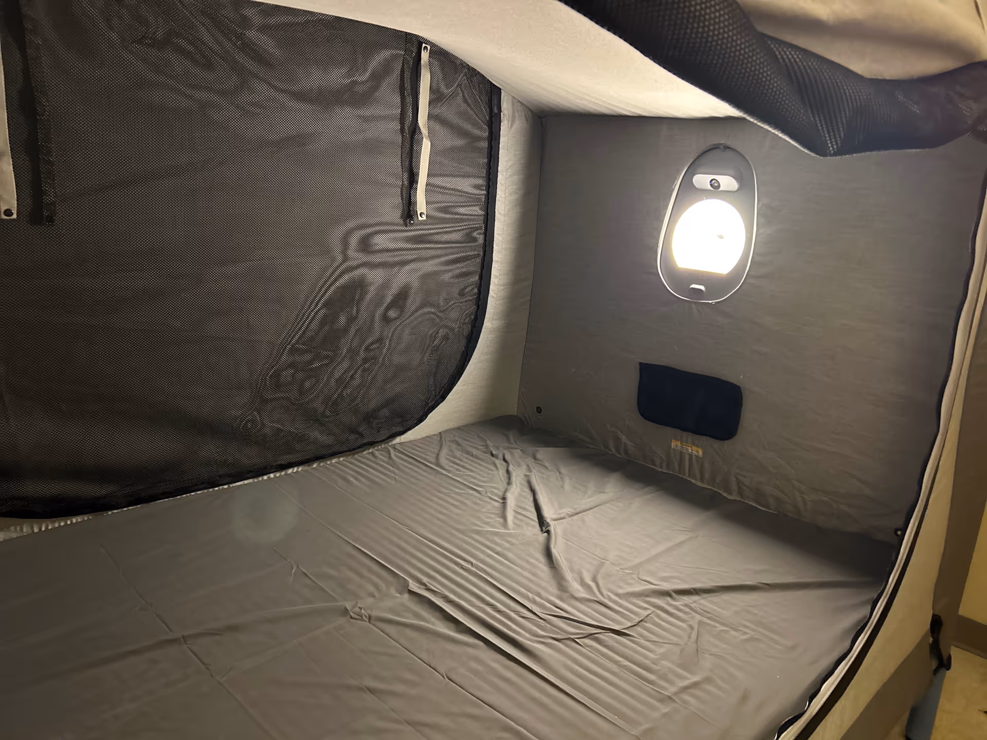 Compact private sleeping pod with a bed, gray sheets, and a wall-mounted light.
