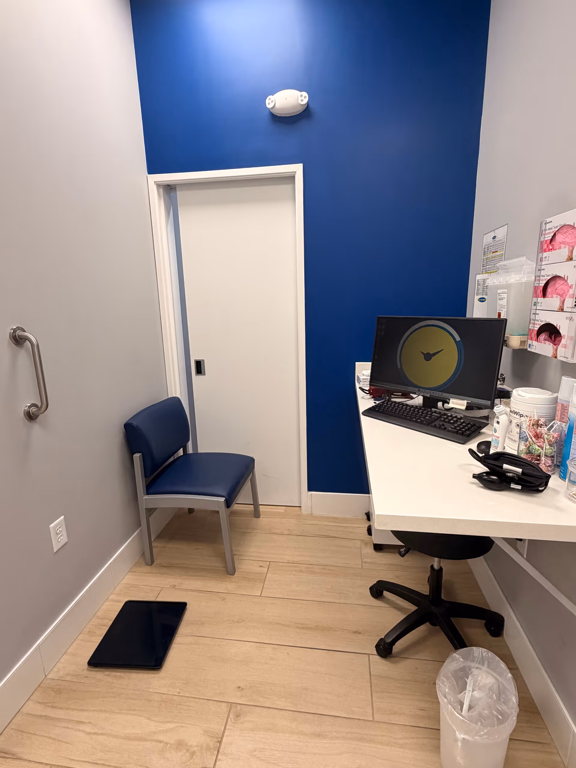 Interior view of clinic-style workspace with blue wall, door, chair, and desk with computer.