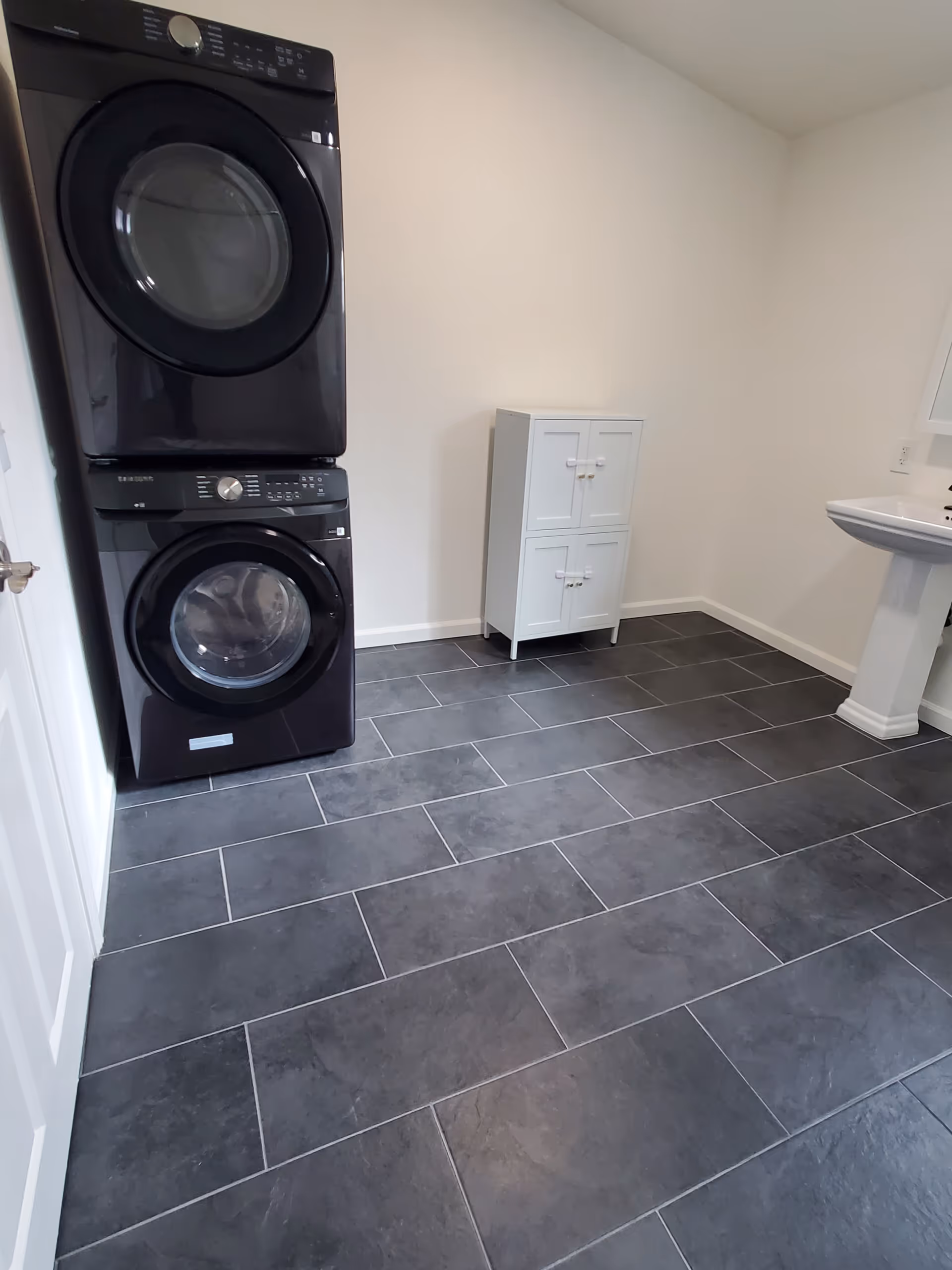Laundry room with stacked washer and dryer, small cabinet, pedestal sink, and tiled floor.