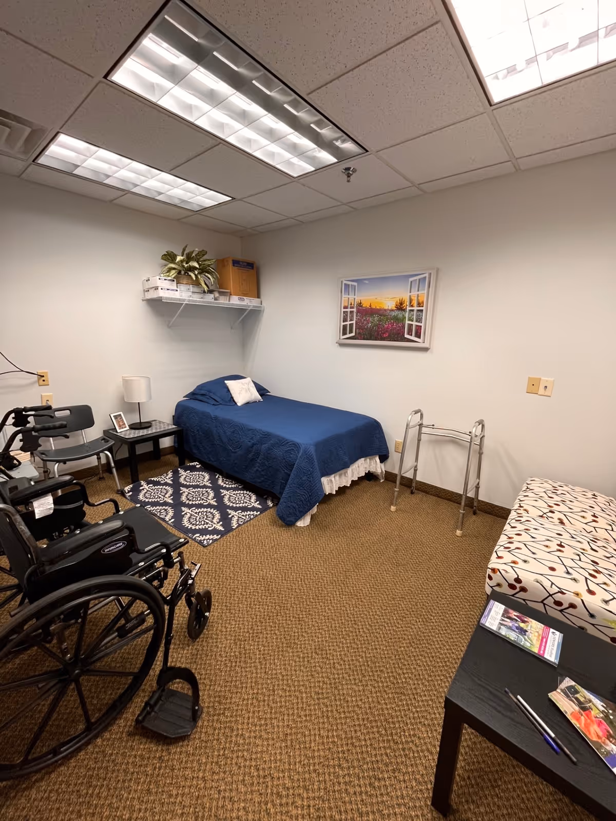 A compact care room with a blue-covered bed, wheelchair, walker, and a patterned rug.