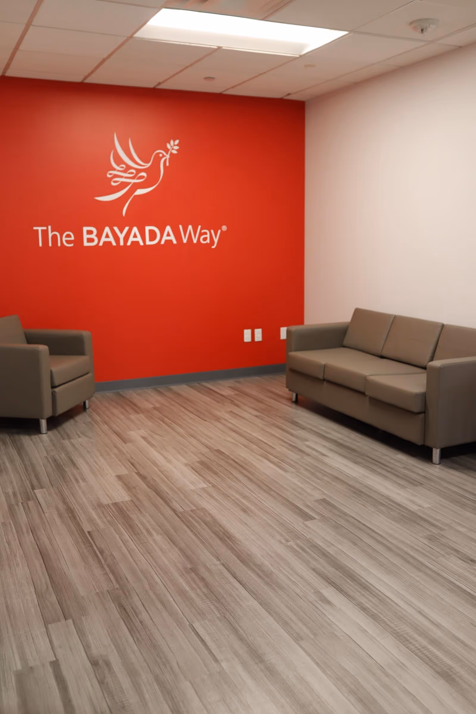 Interior lounge with beige couches and a red wall featuring The BAYADA Way logo.