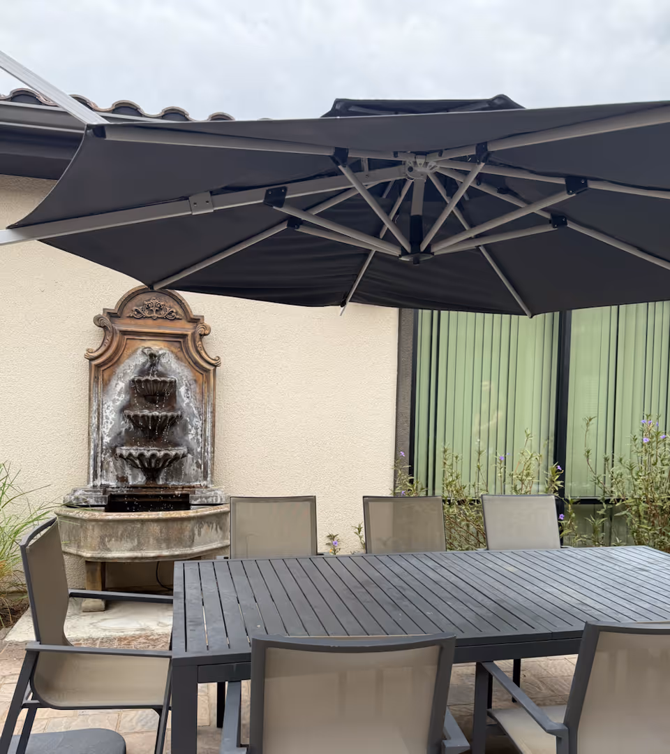 Outdoor patio with a large umbrella, dining table, chairs, and a fountain against a building.