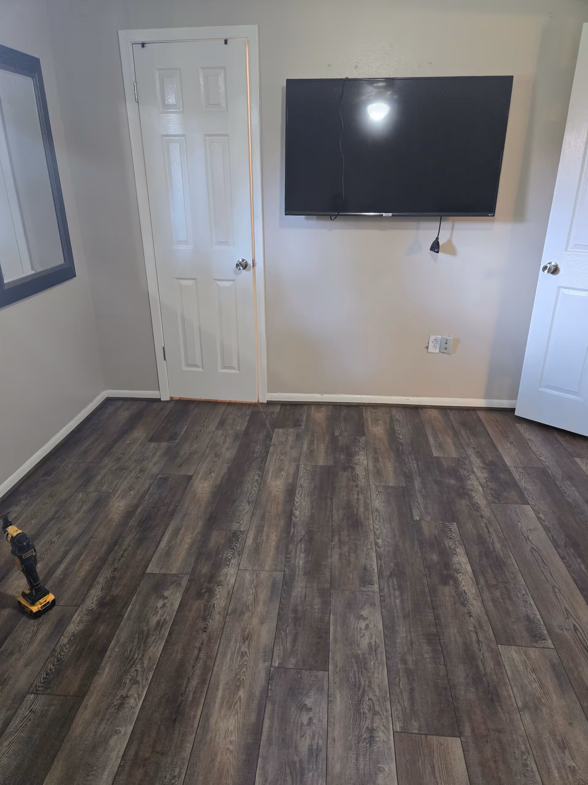 Empty room with wood-like flooring, wall-mounted TV, two doors, and a drill on the floor.