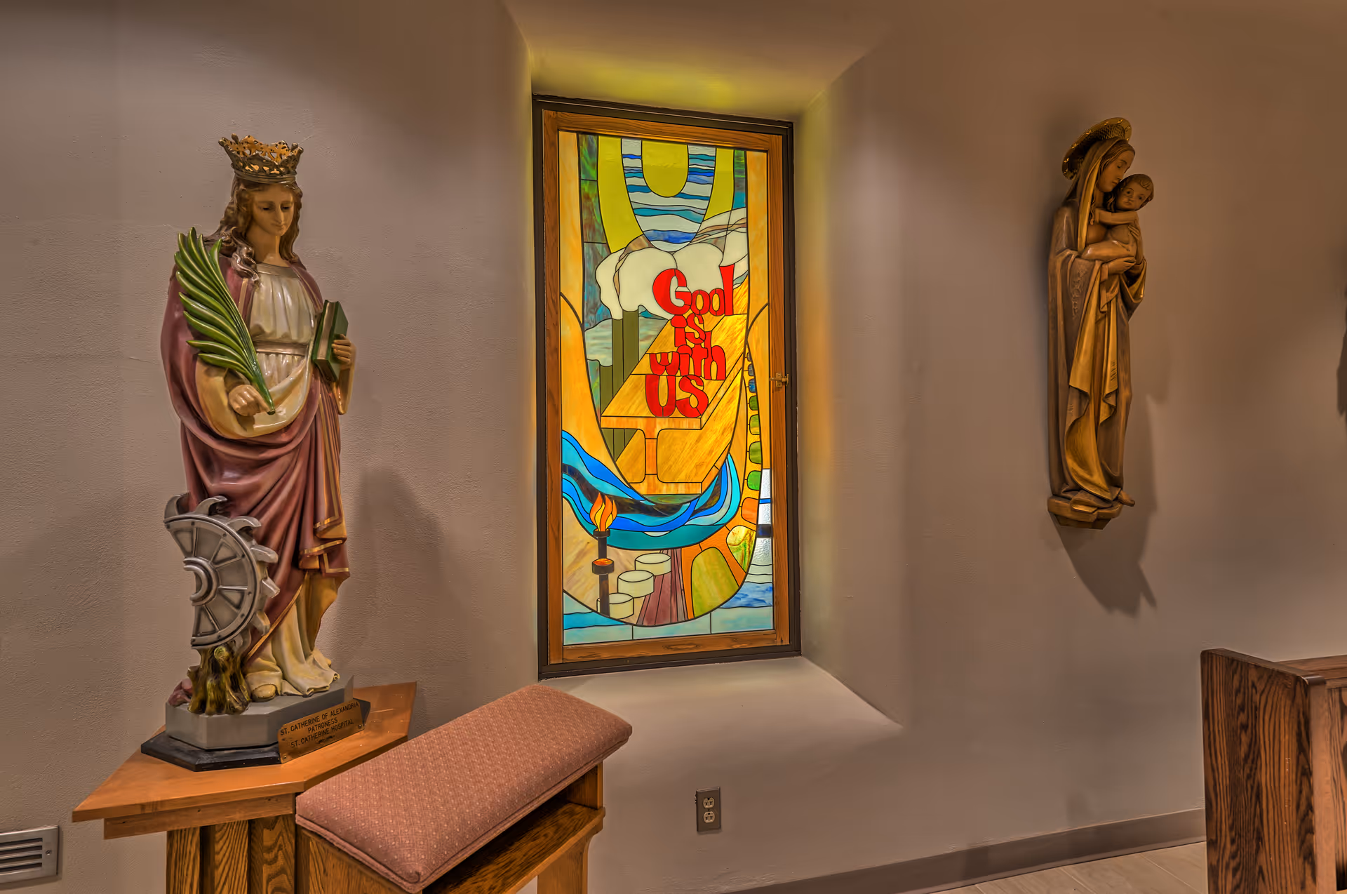 Interior hallway featuring statues and a stained-glass window with the message God is with us.