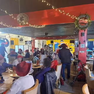 Busy dining hall with seniors seated at tables, caregivers mingle under festive string lights.