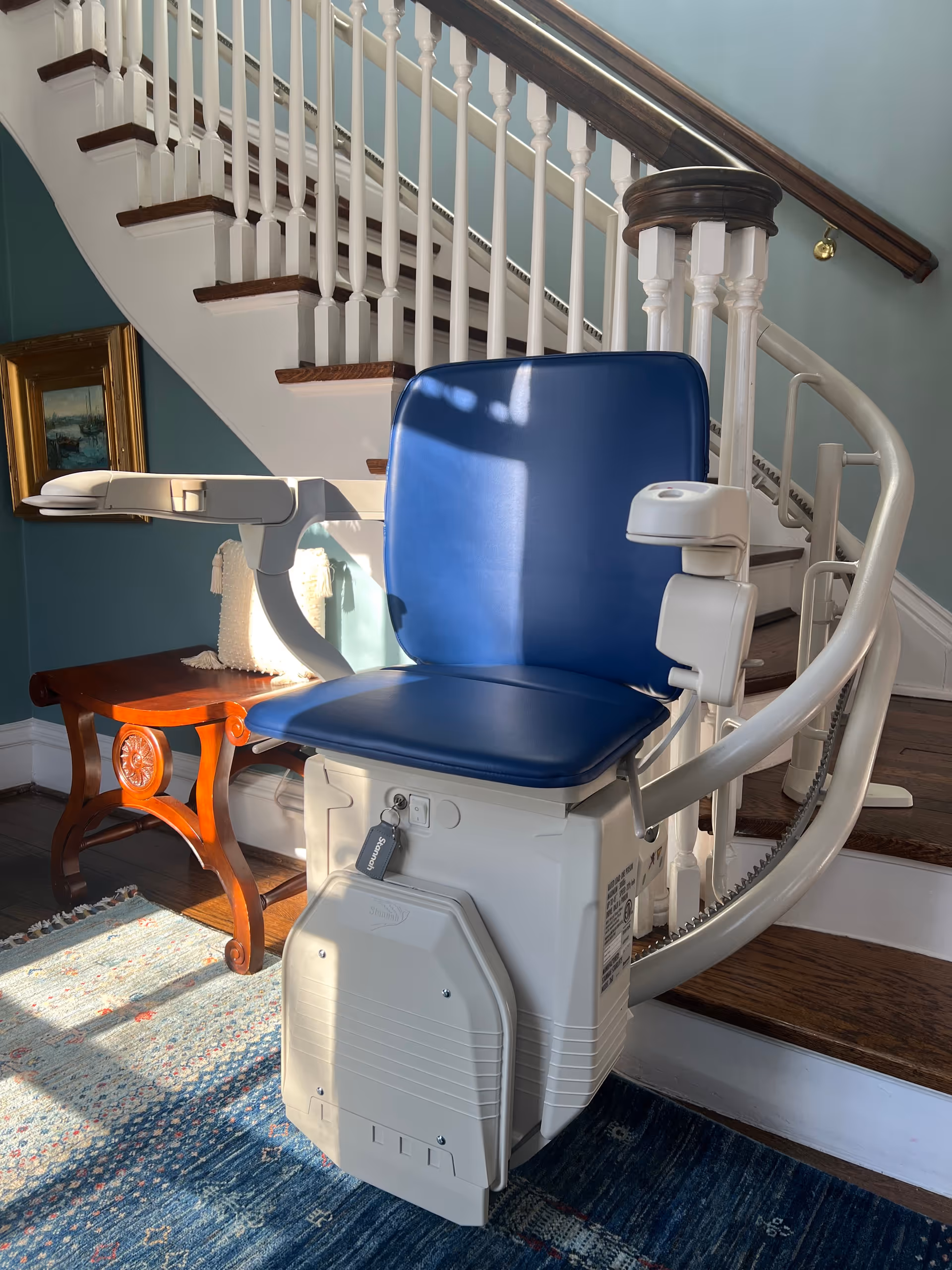 Blue stairlift chair mounted on a curved indoor staircase beside a wooden side table.