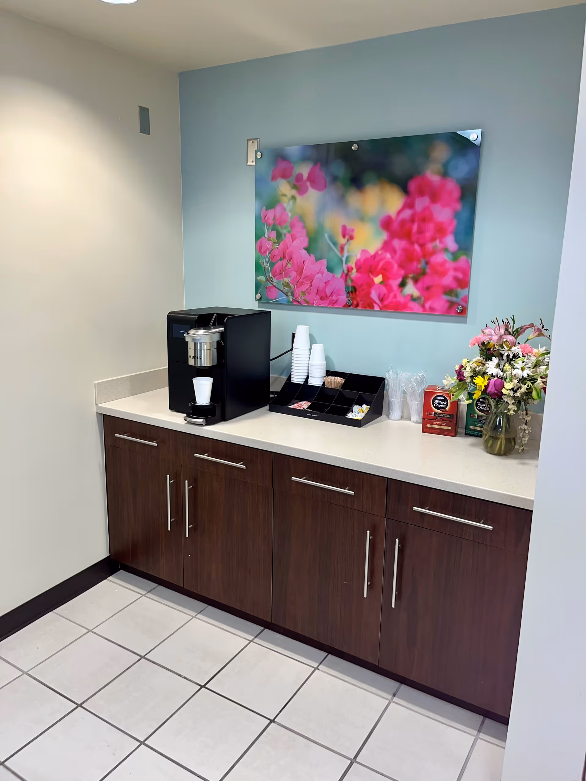 A beverage station with coffee machine, cups, and a flower arrangement along a blue wall.