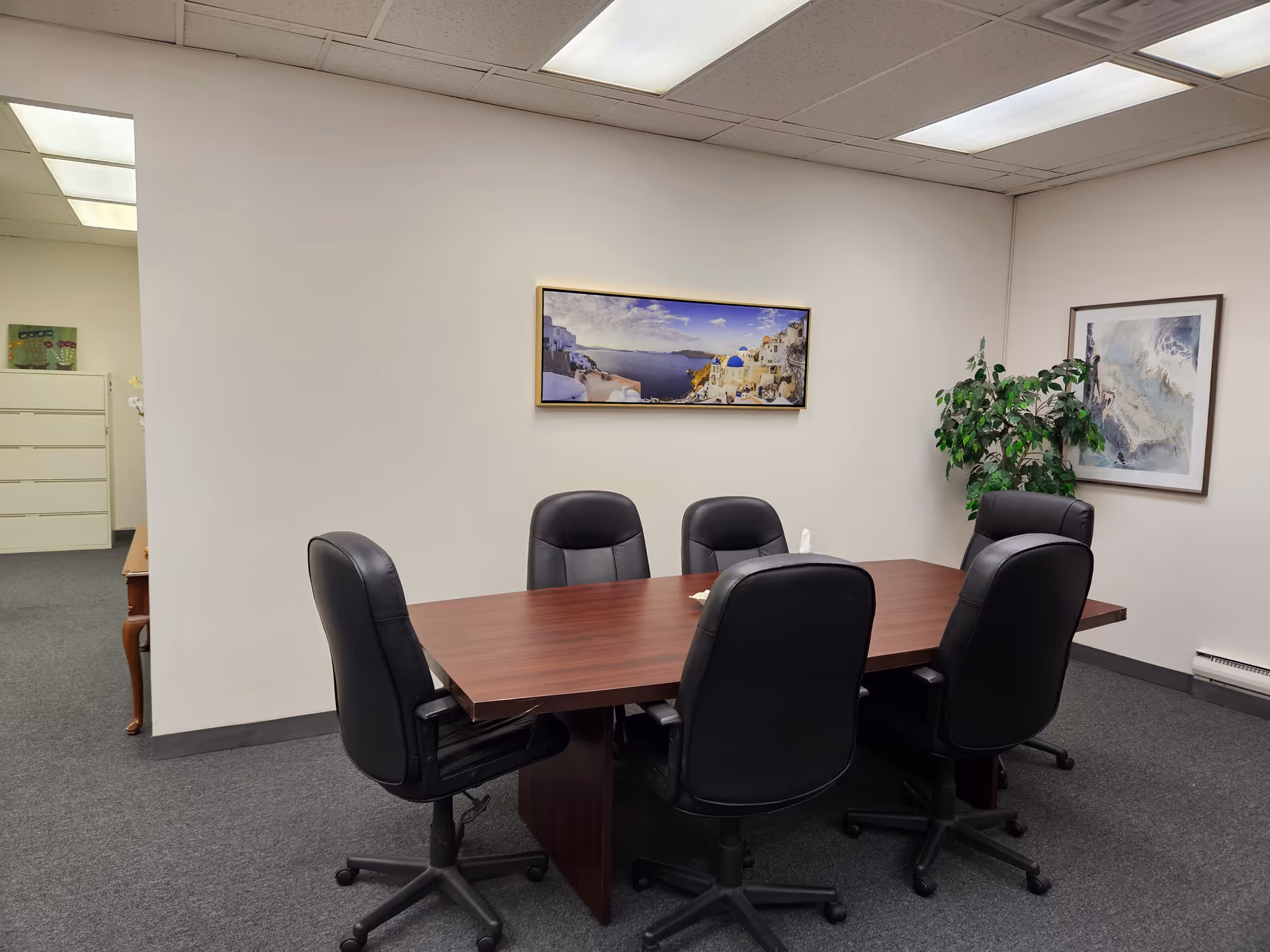 A conference room with a wooden table, six black chairs, plant, and wall art.