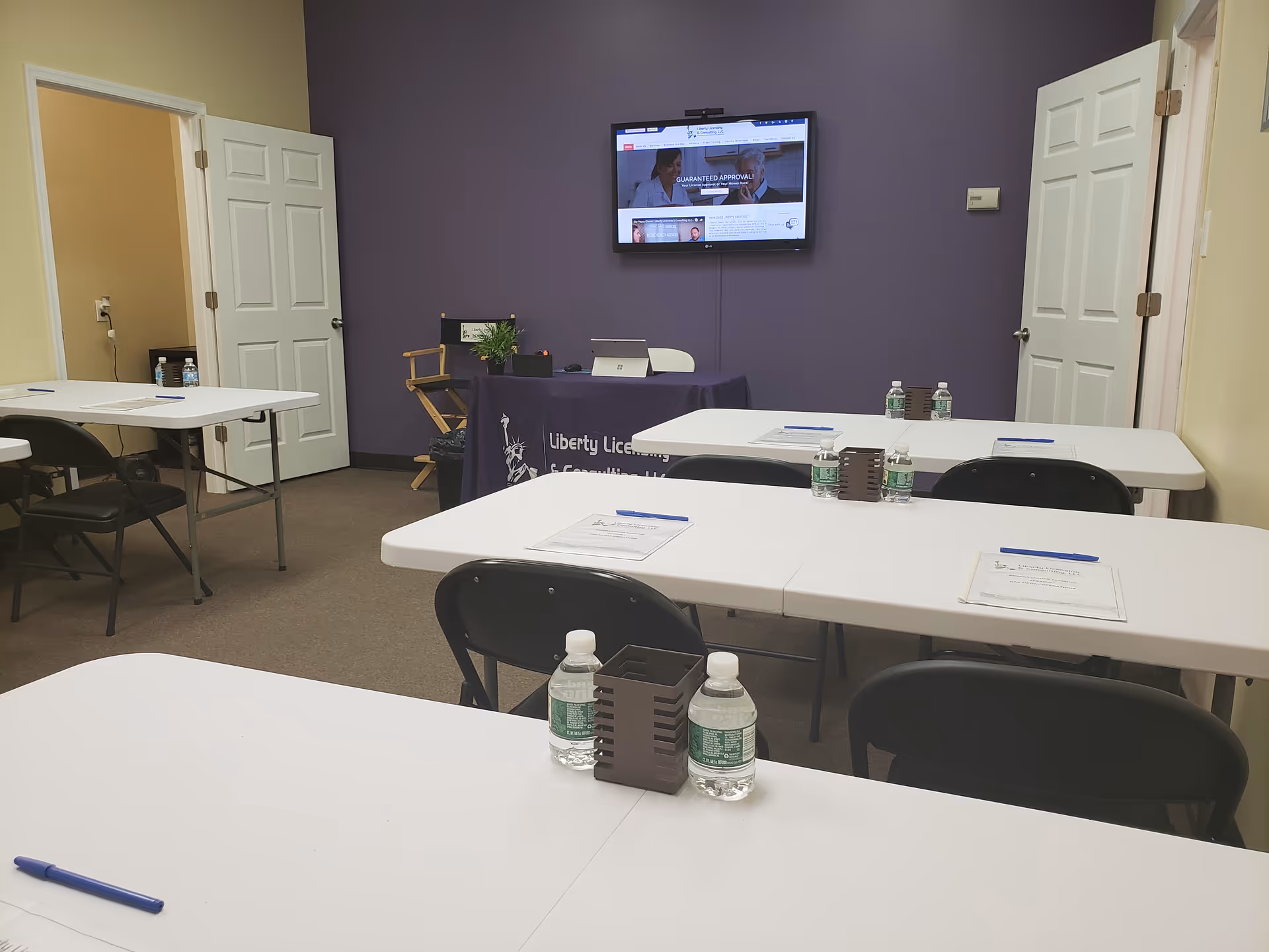 Conference room setup with white tables, black chairs, water bottles, and a mounted screen.