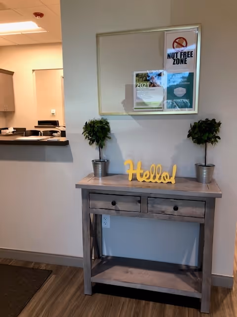 Wood console table with two potted plants, Hello sign, and bulletin board in a hallway.