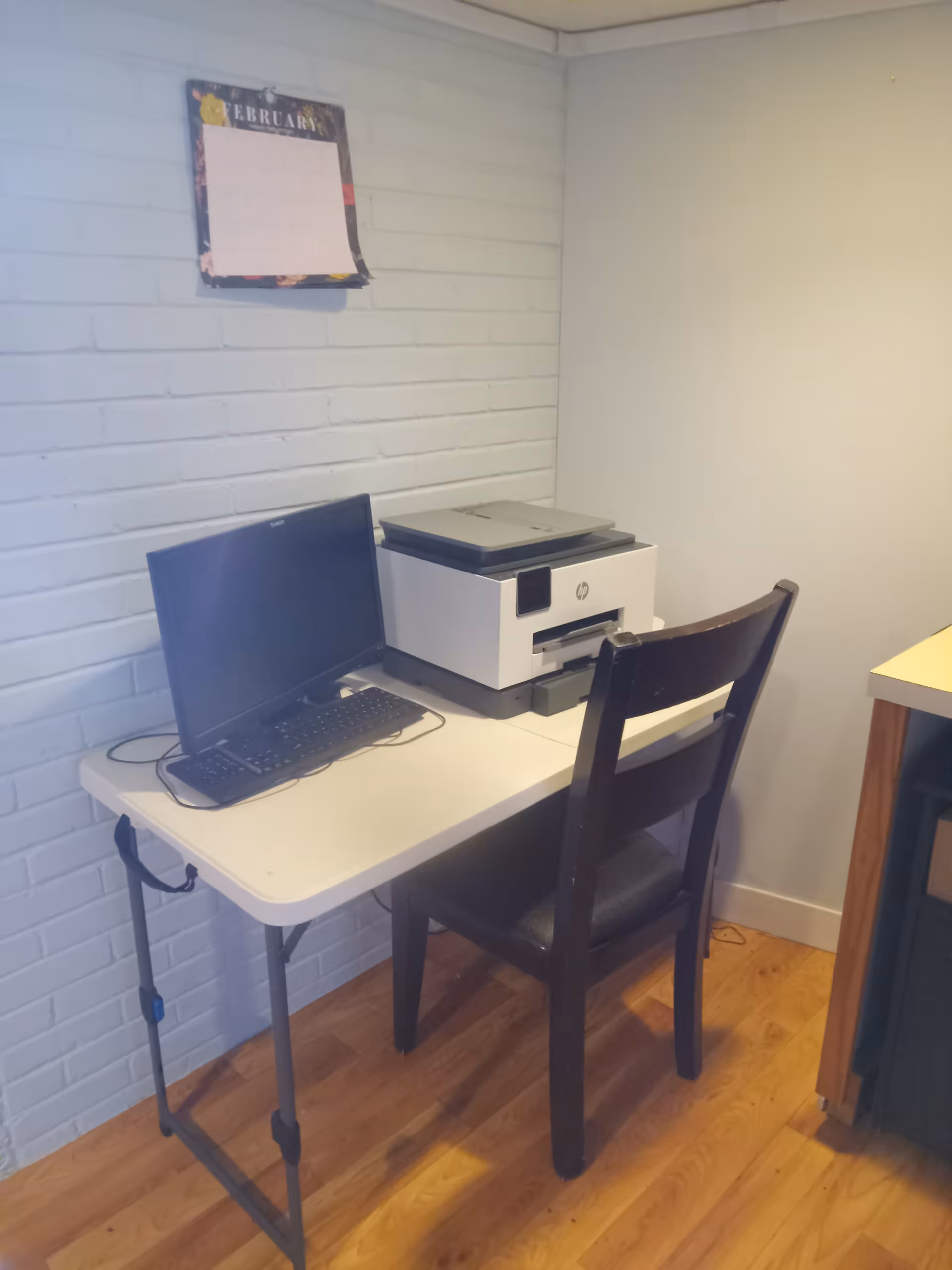 Small desk with computer, printer, and calendar against a light blue brick wall.