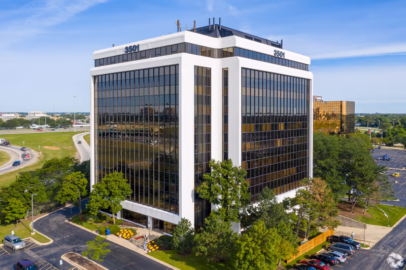Aerial view of a modern glass-front office building surrounded by trees and parking.