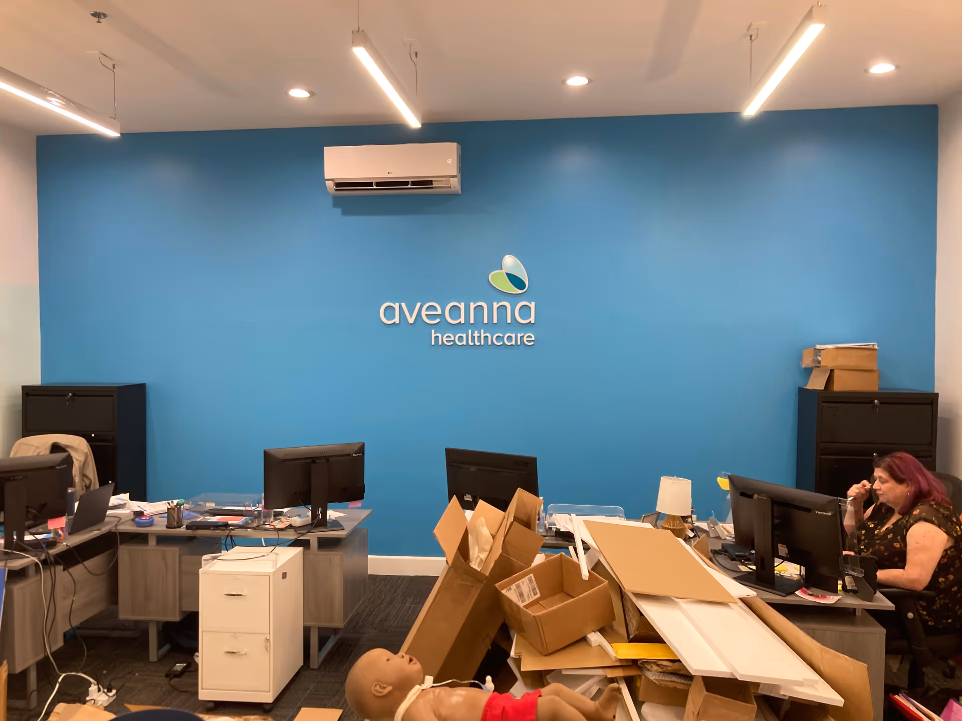 Blue wall with Aveanna Healthcare logo behind cluttered desks and a worker at a computer.