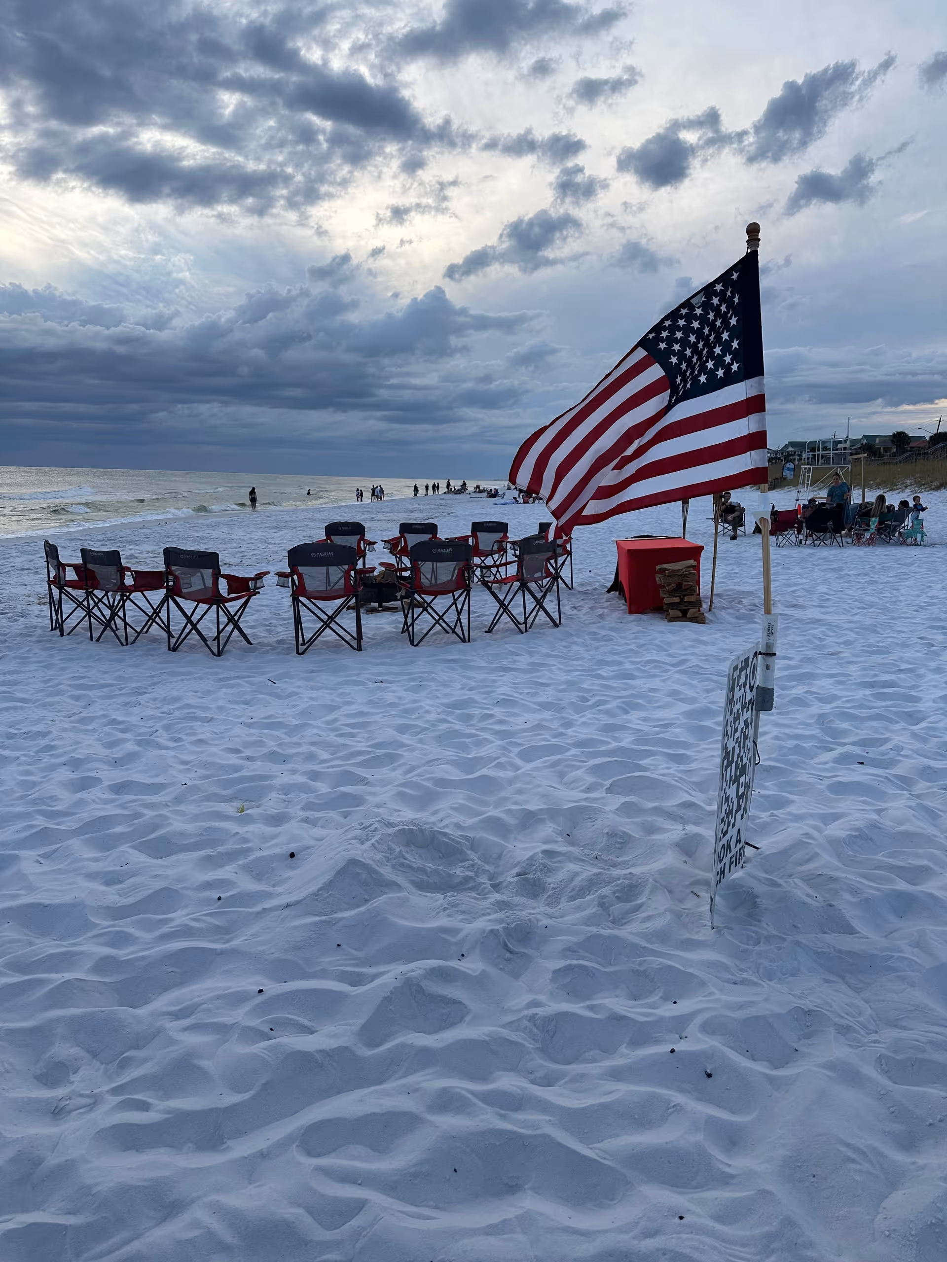 Outdoor beach gathering with folding chairs, an American flag, and people near the shoreline.