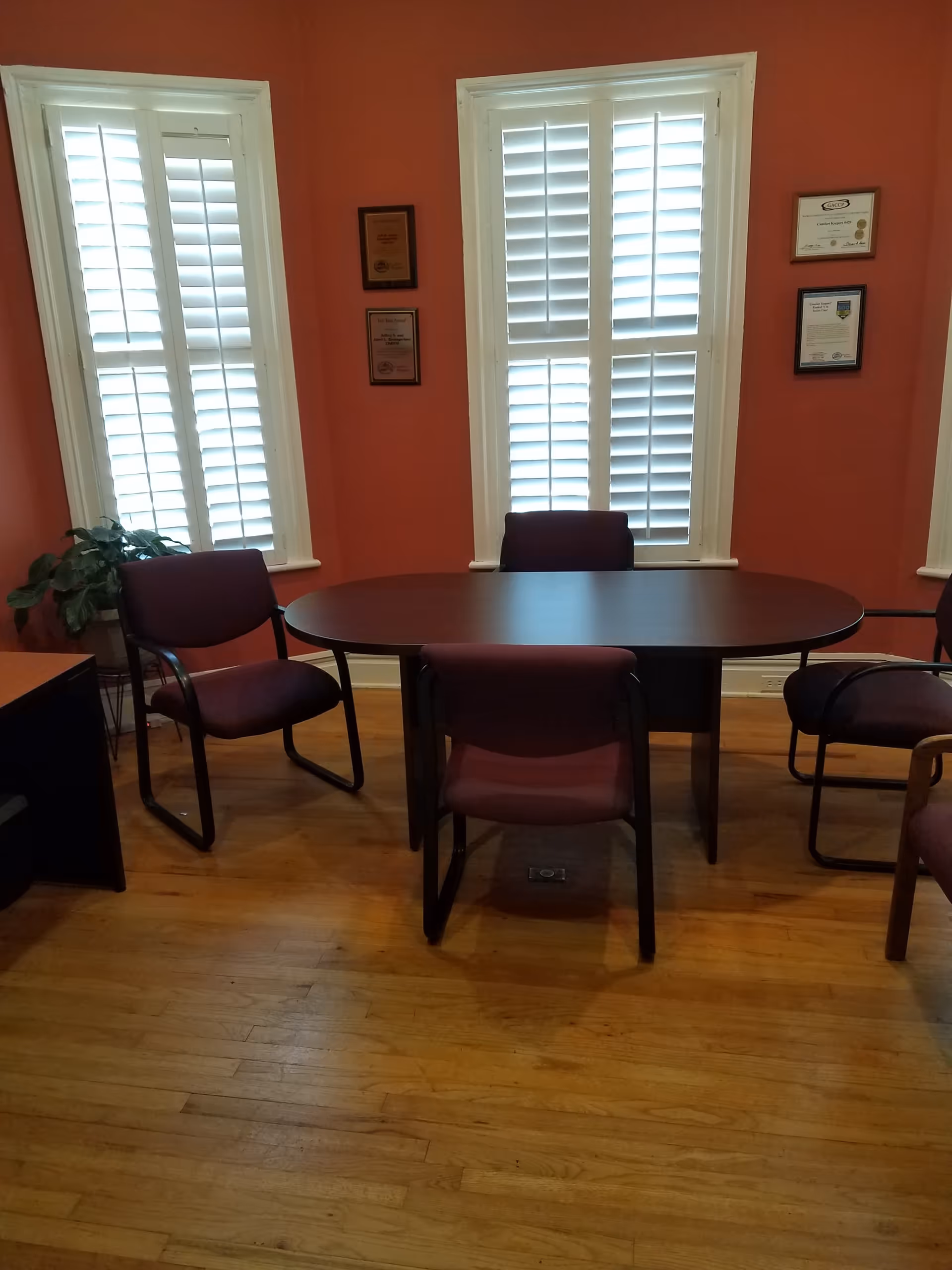 An oval table with maroon chairs in a pink dining room with window shutters.