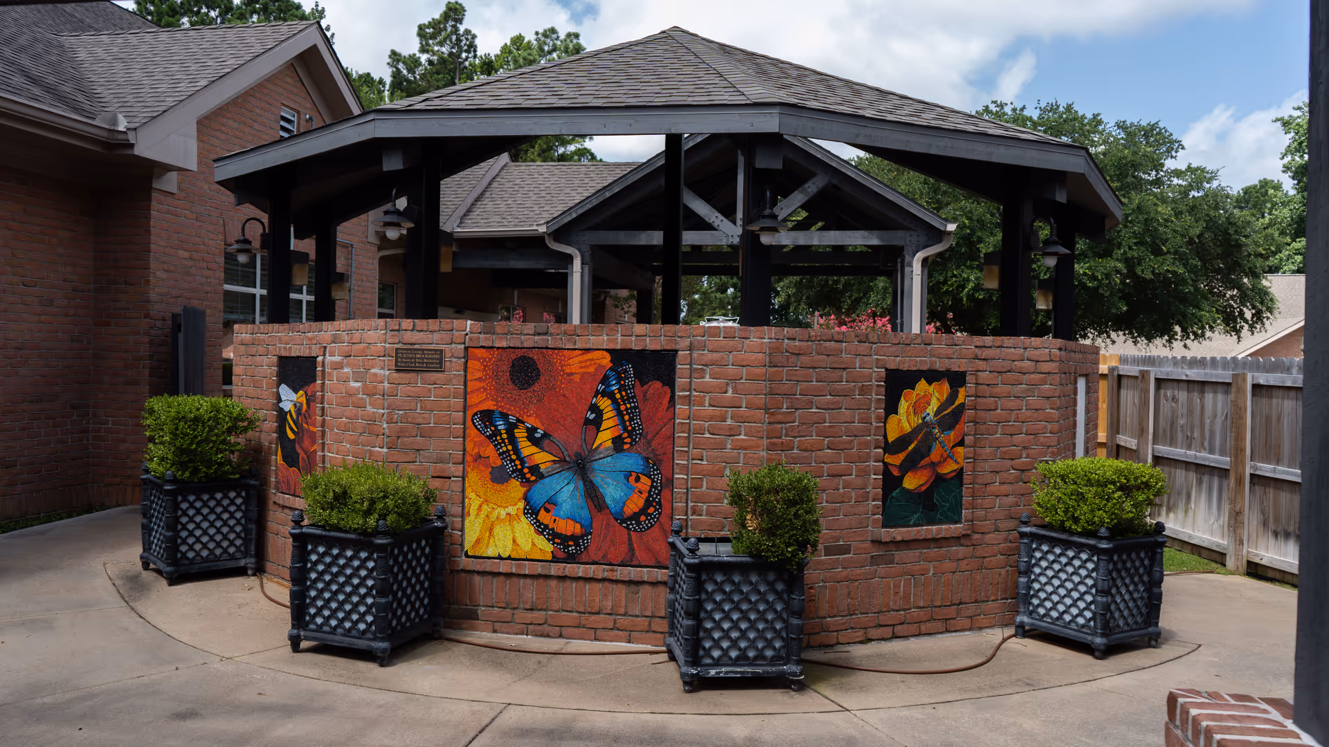Brick outdoor pavilion in a courtyard with butterfly artwork and potted shrubs.