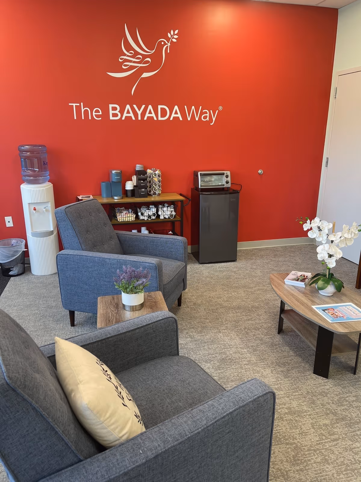 Red-walled reception area with BAYADA logo, gray chairs, coffee station, and plants.