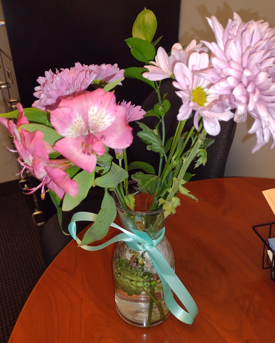 Vase of pink flowers with green leaves sits on a wooden round table indoors.