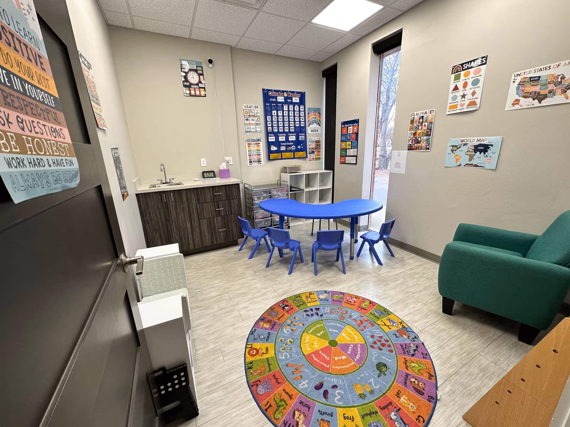 Indoor therapy room with blue table and chairs, circular colorful rug, kitchenette area, and green sofa.