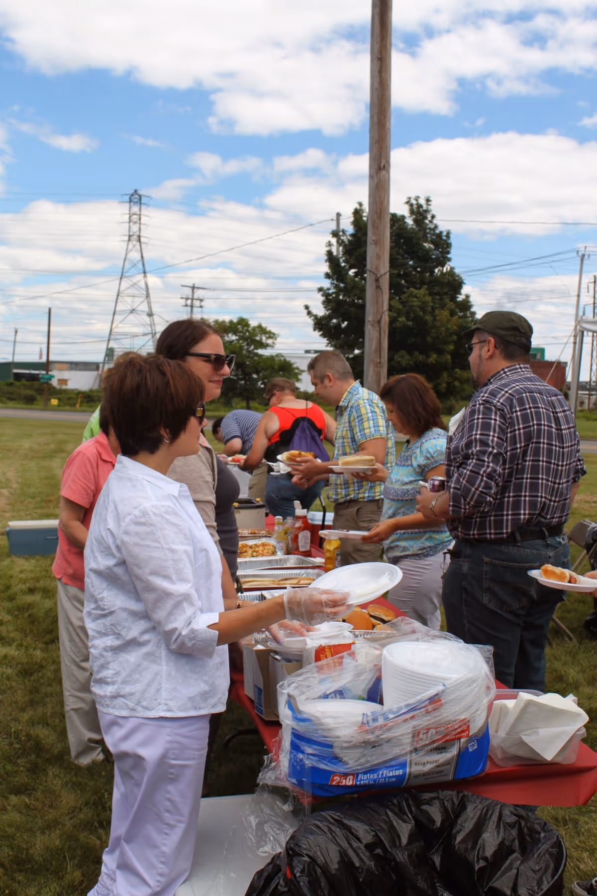 Outdoor gathering of people around a table with food and plates.